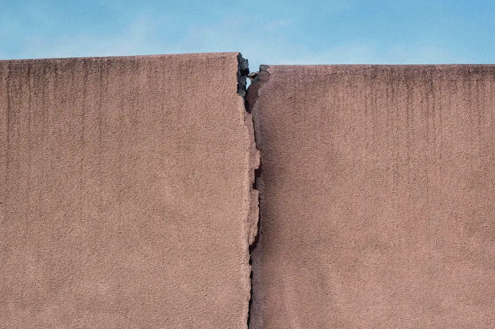 Abstract photograph of a deep vertical crack in a textured concrete wall against a clear blue sky, reflecting the tension between structural imperfection and the persistence of creation.