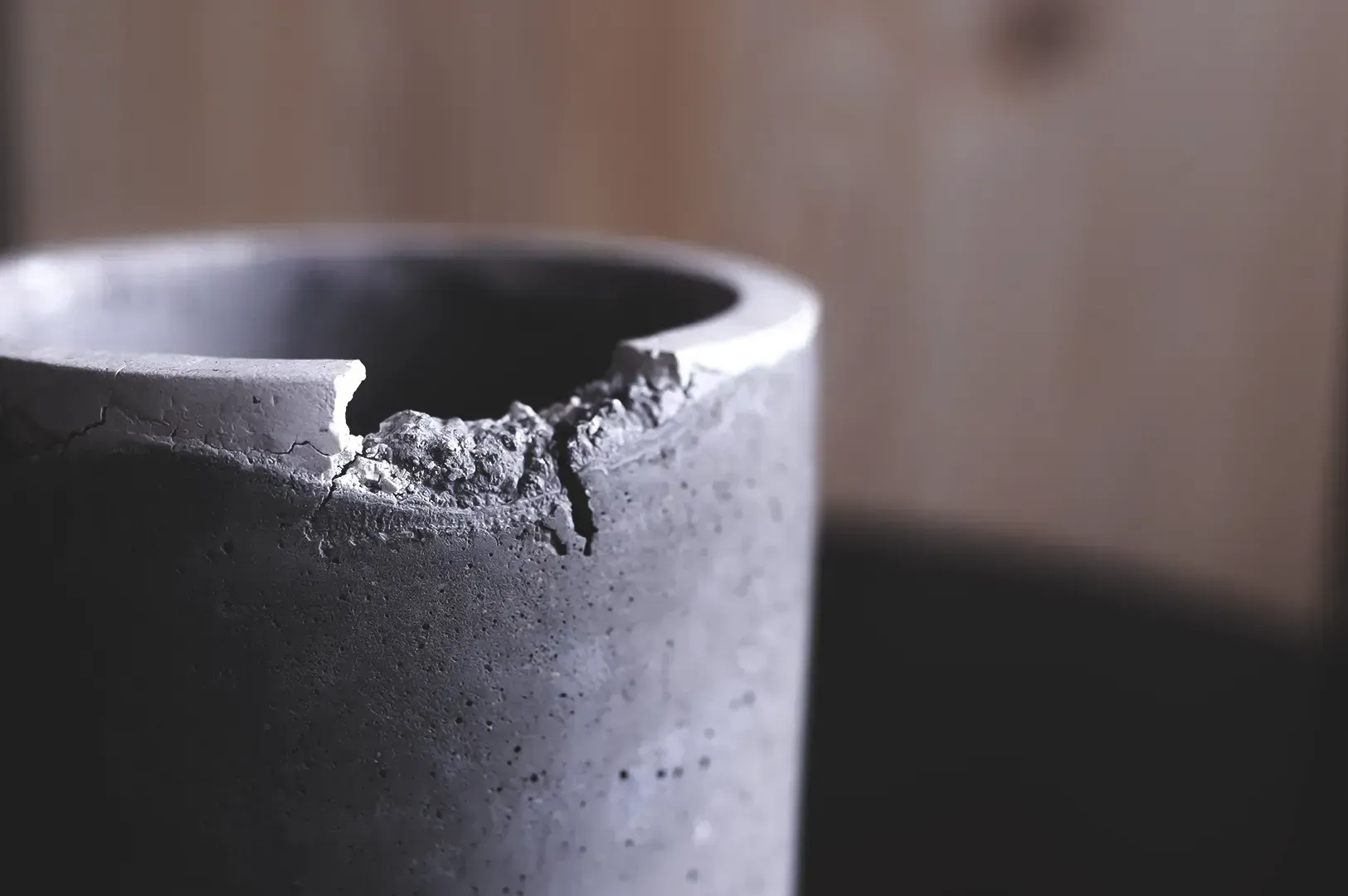Close-up shot of a minimalist grey concrete pot with a chipped rim resting against a warm wooden backdrop, capturing the raw beauty of worn objects that resist modern discard culture.