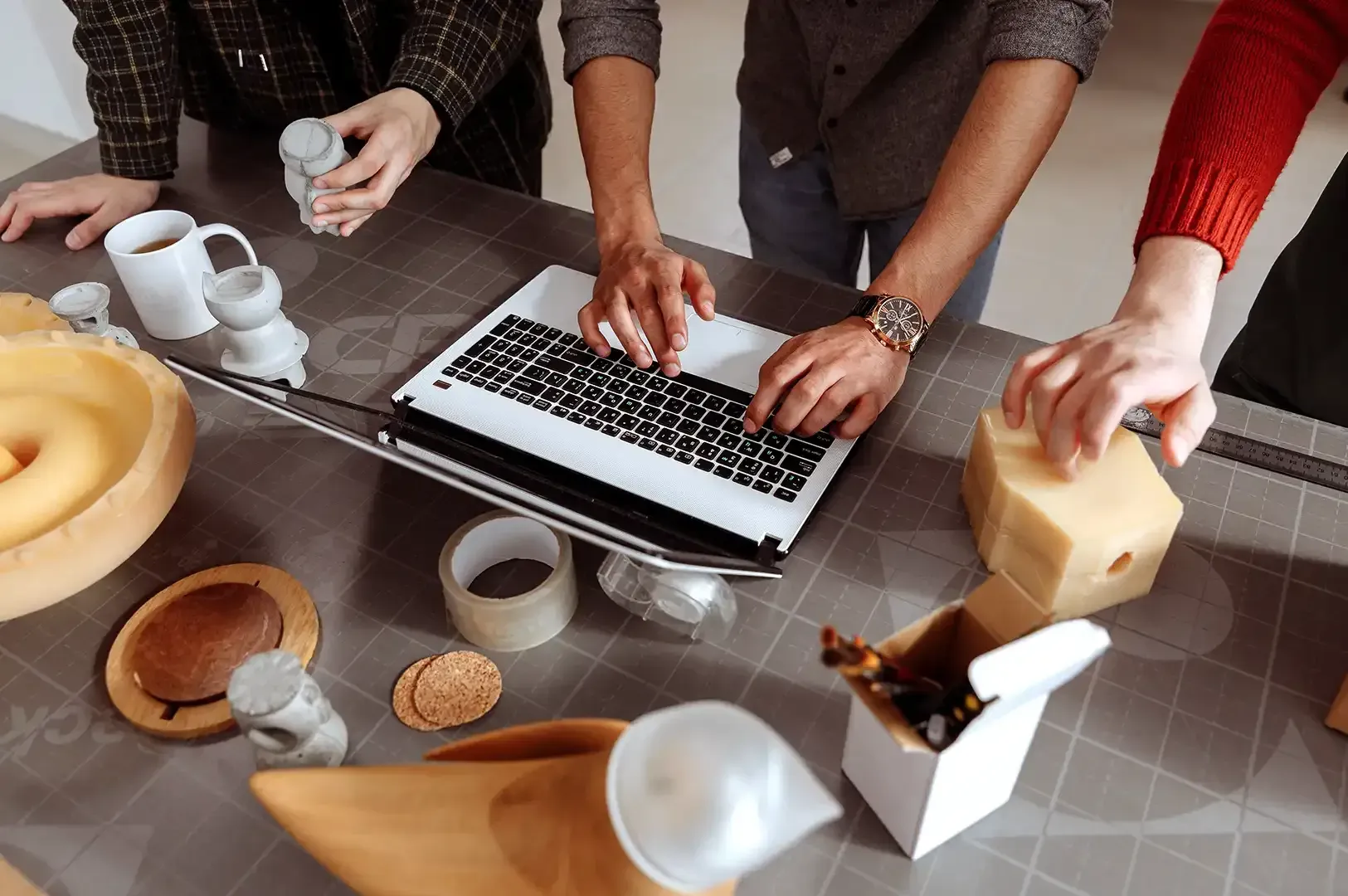 A collaborative team of designers working around a laptop alongside 3D prototypes, wooden crafts, and materials, illustrating how craftsmanship and shared standards strengthen modern business systems.