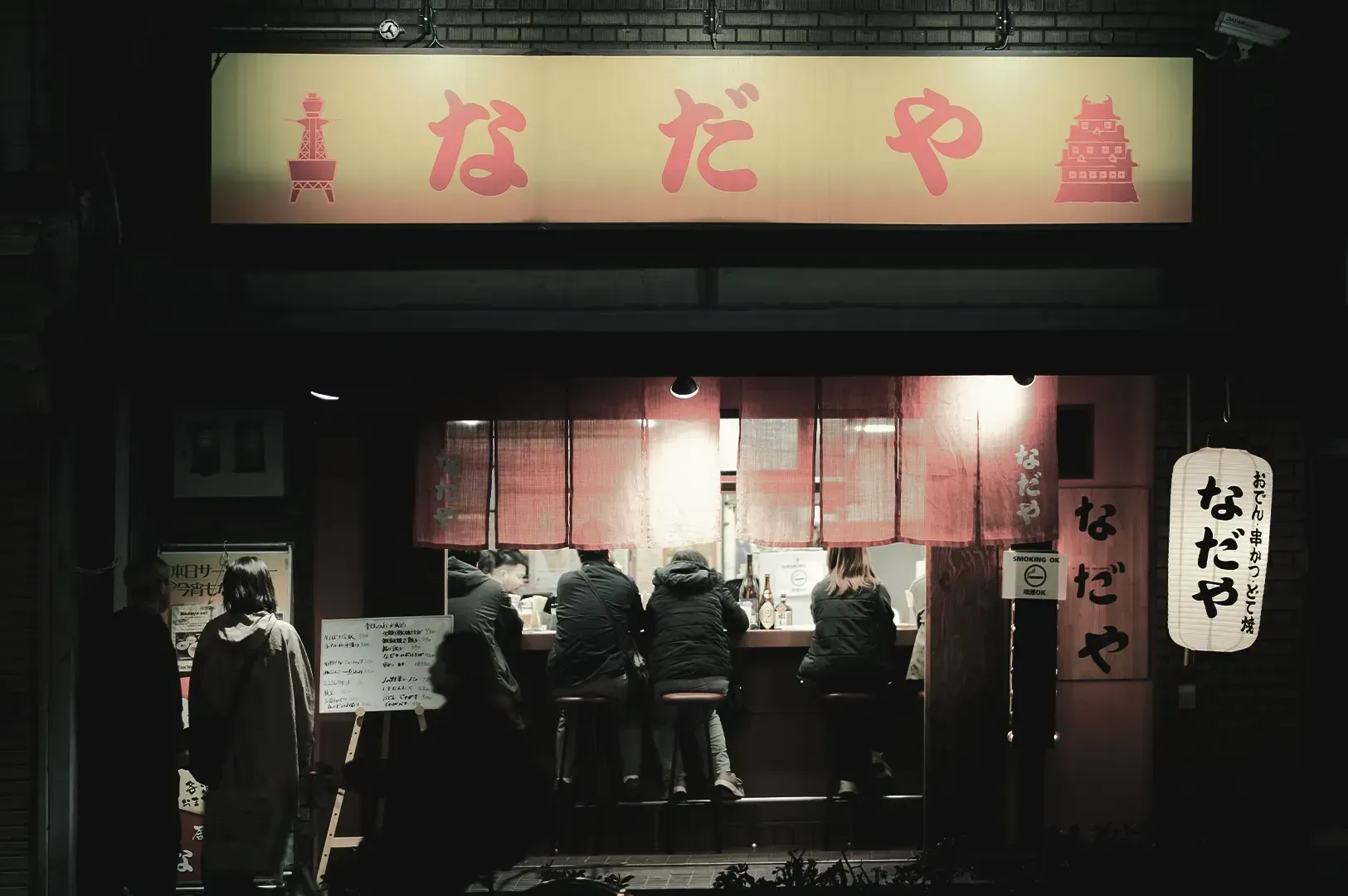 A cozy Japanese izakaya at night with patrons seated at the counter, highlighting how local sounds and social spaces turn strangers into a sense of belonging.