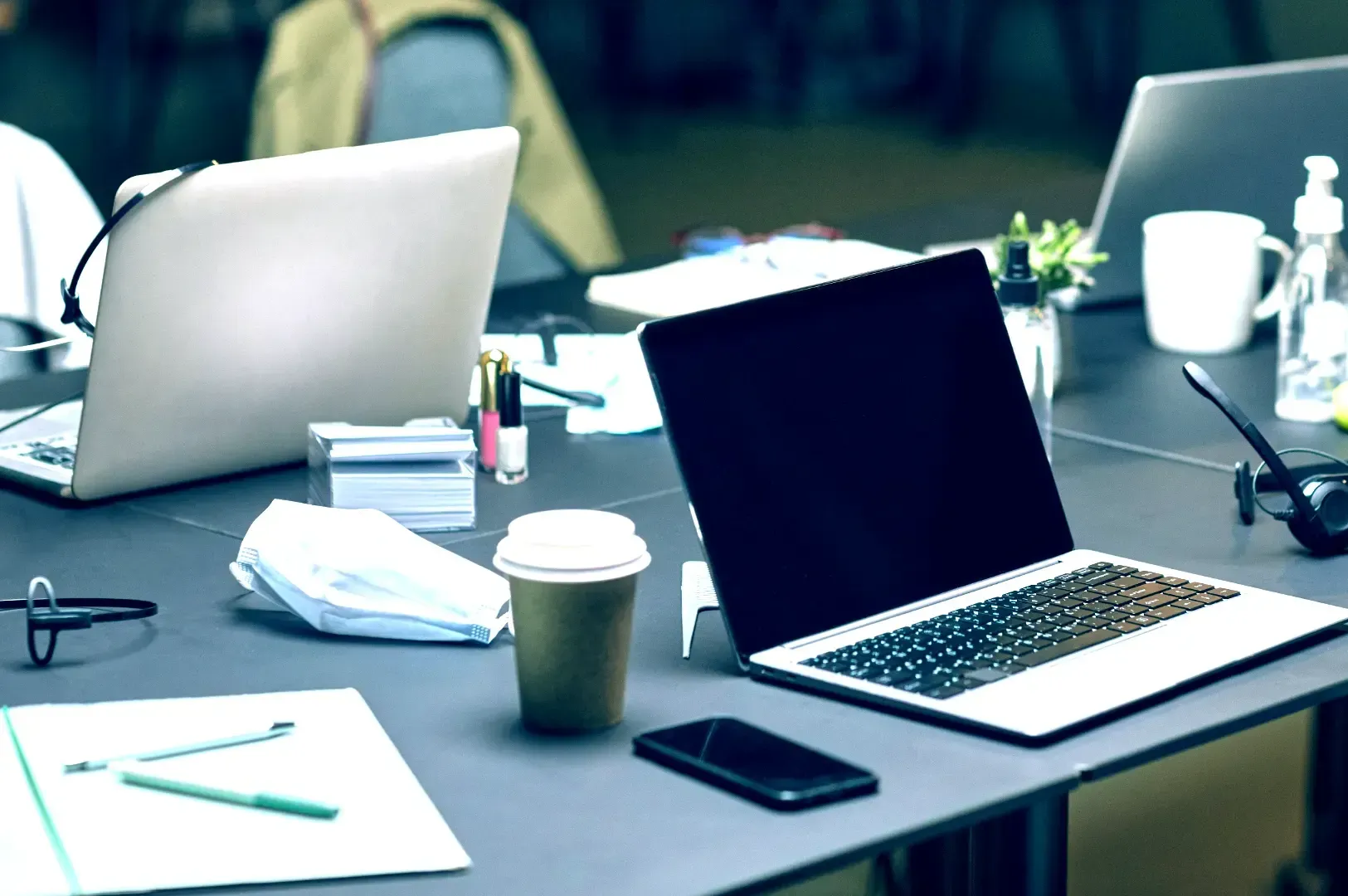 A cluttered shared office desk filled with open laptops, headsets, coffee cups, and papers, highlighting everyday work habits and workplace standards.