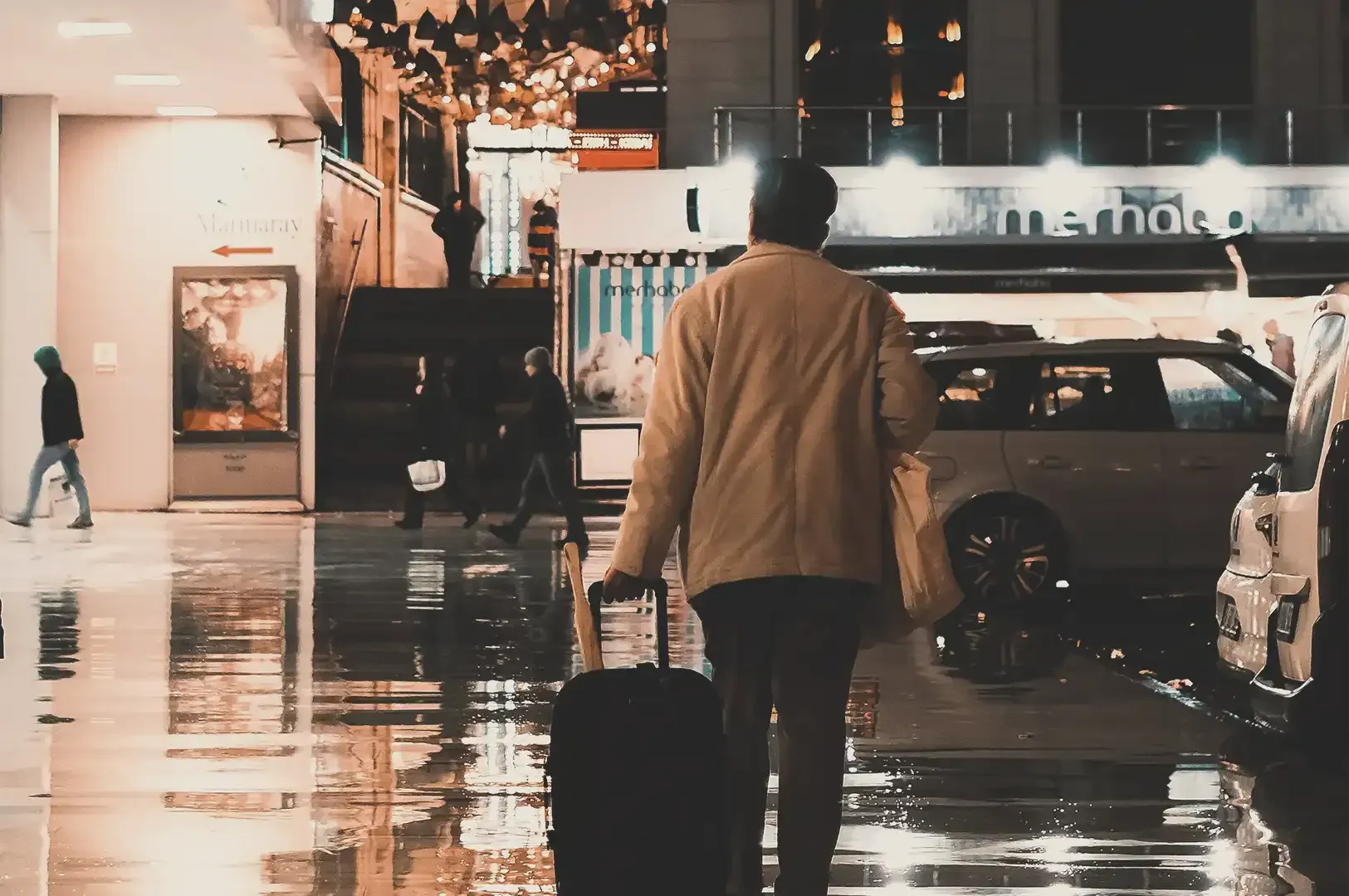 A traveler walking down a wet city street at night, pulling a small suitcase and carrying a light bag, highlighting the ease of minimalist travel through urban environments.