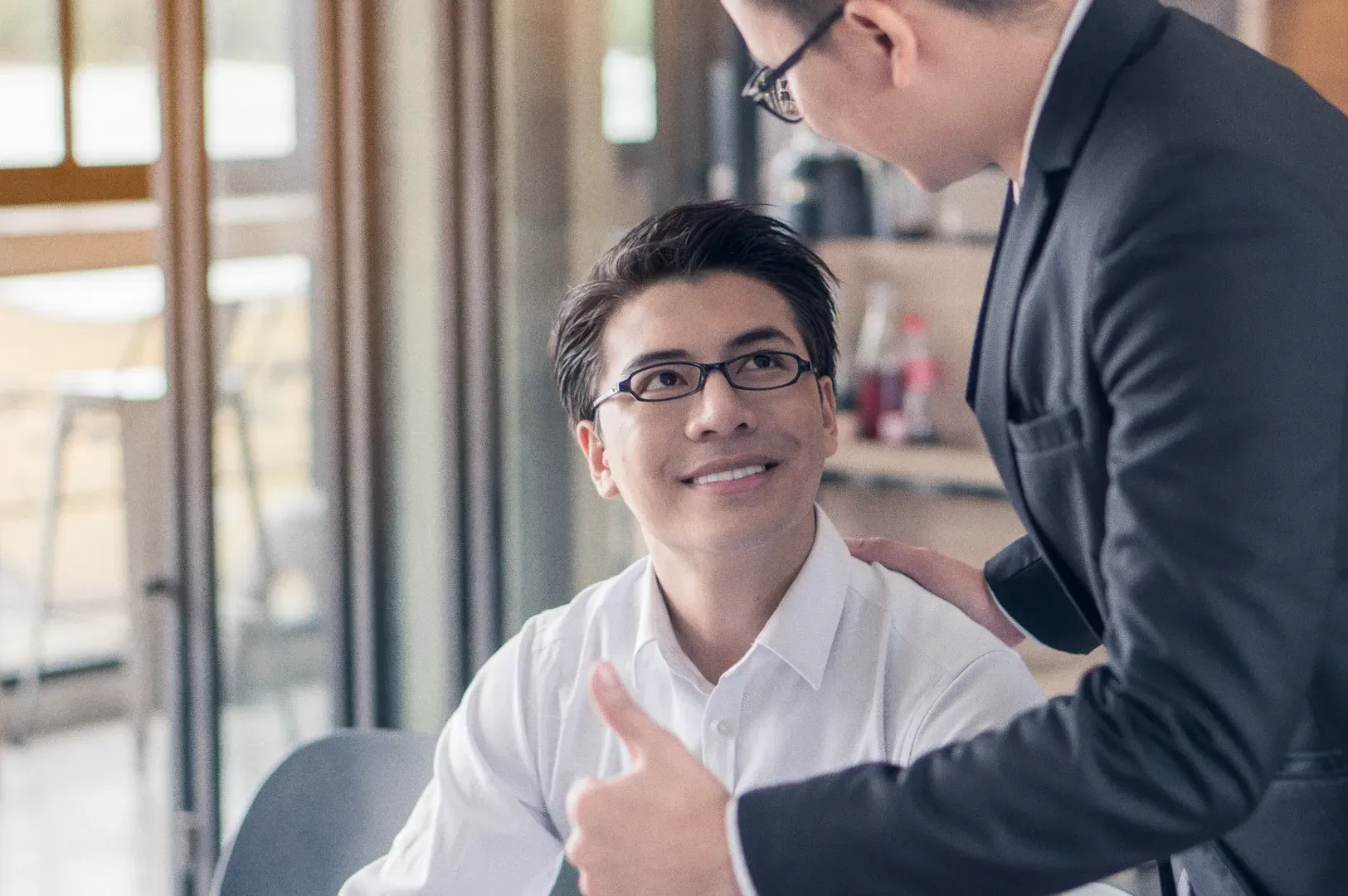 Eye-level medium shot of two professionals seated at a desk in an office, with one person gesturing while discussing work tasks.