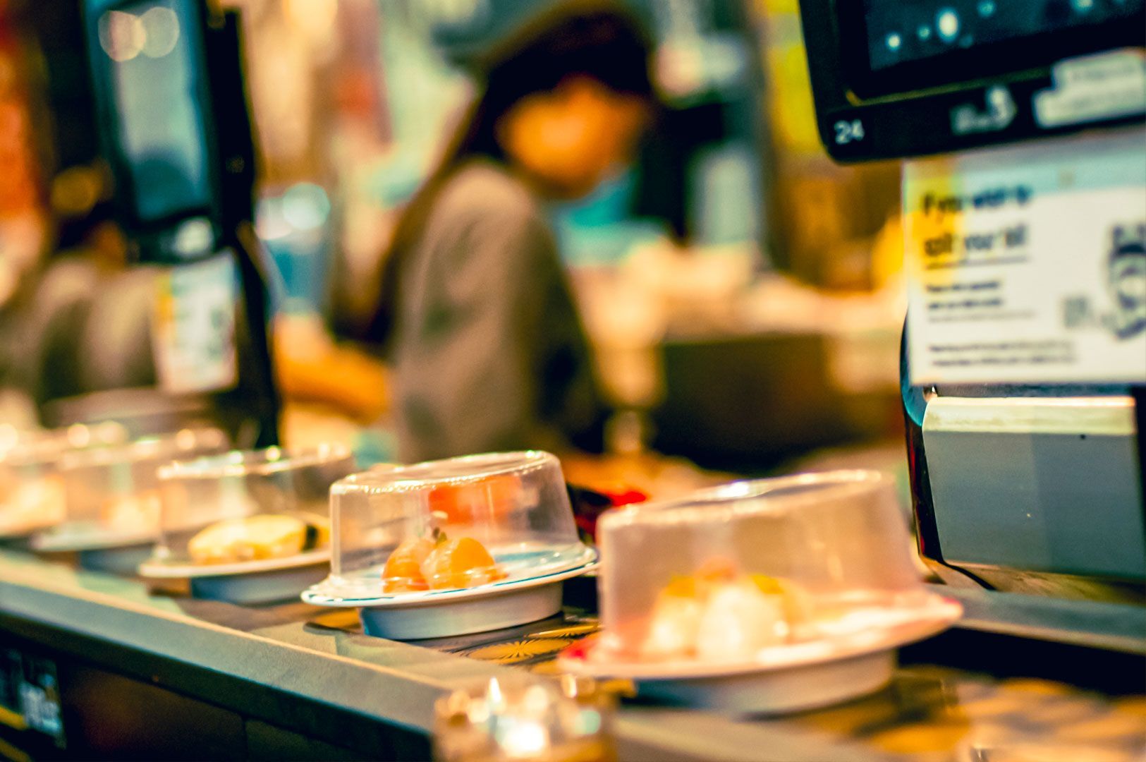 Plates of sushi covered with clear domes moving along a conveyor belt inside a modern Japanese sushi restaurant.