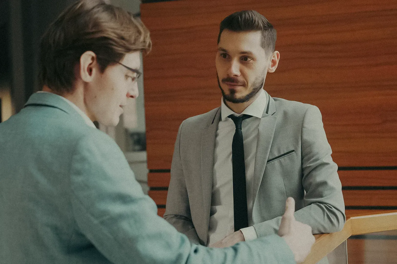 Conversation between two professionals standing near a wooden railing in a corporate office interior.