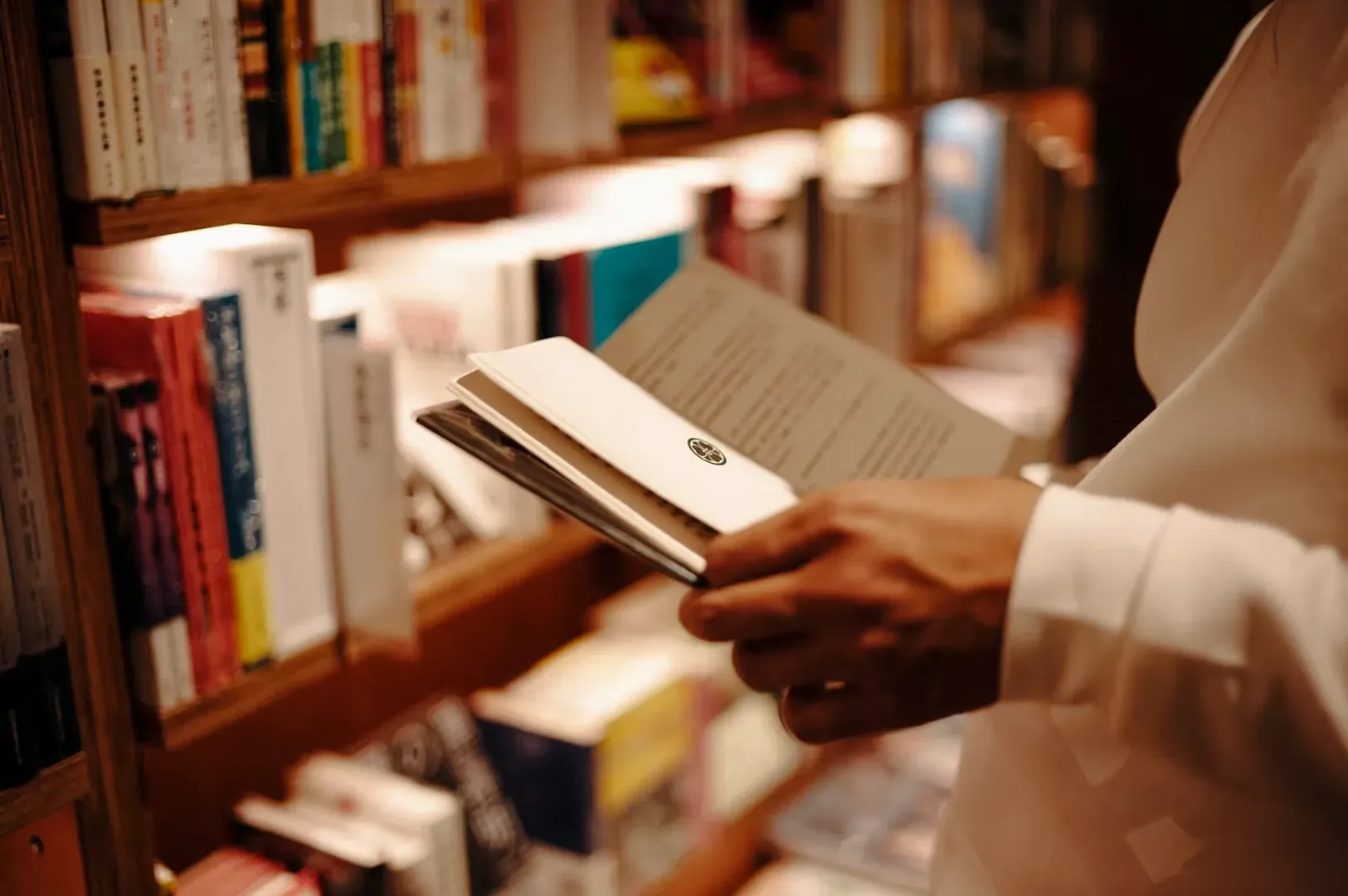 Close‑up of hands holding an open book inside a warmly lit bookstore with shelves lined with various titles.