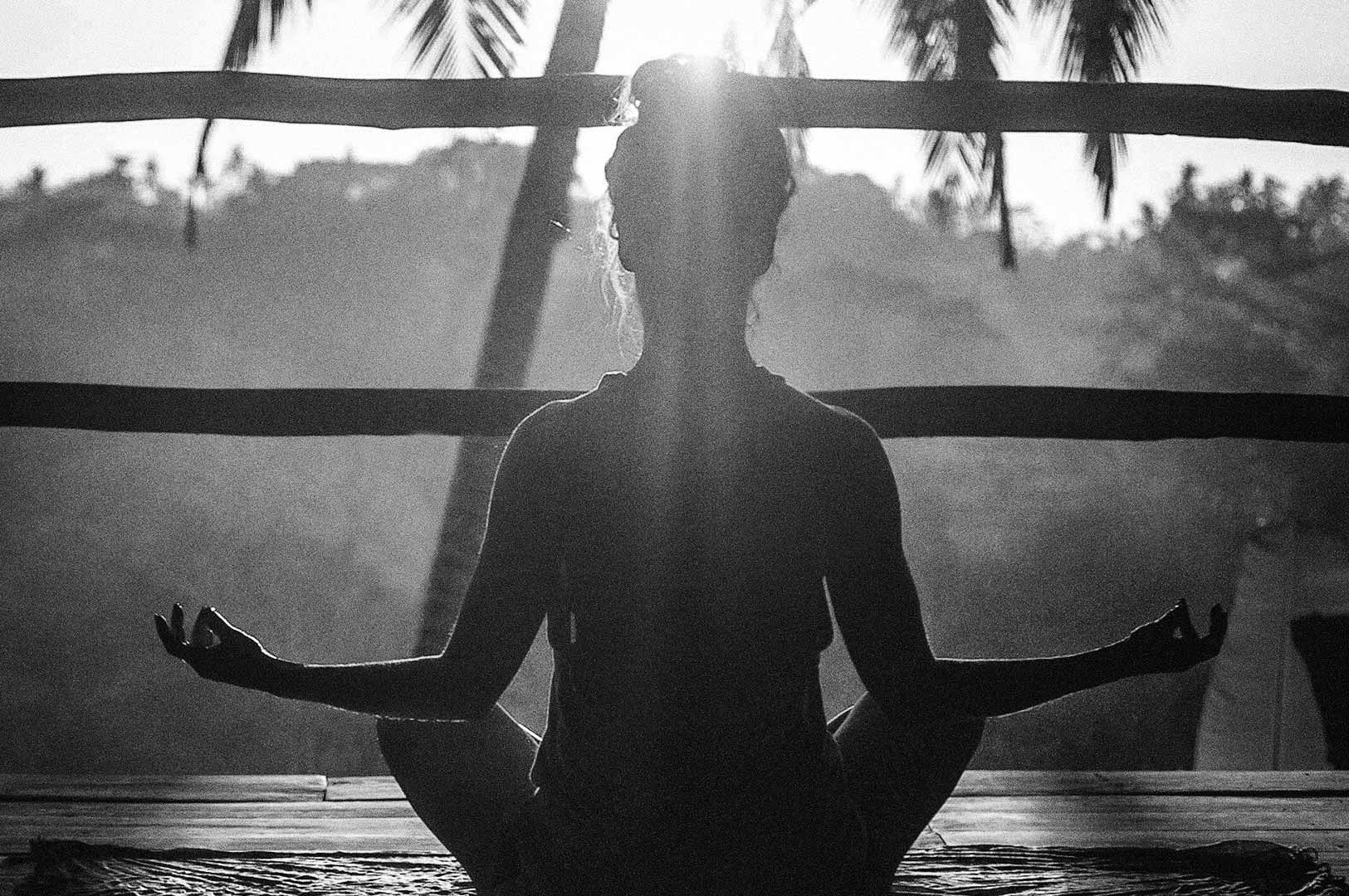 Silhouette of a person practicing yoga in lotus pose on a wooden deck, seated cross‑legged with hands in a mudra, palm trees and sunlight streaming through the background.