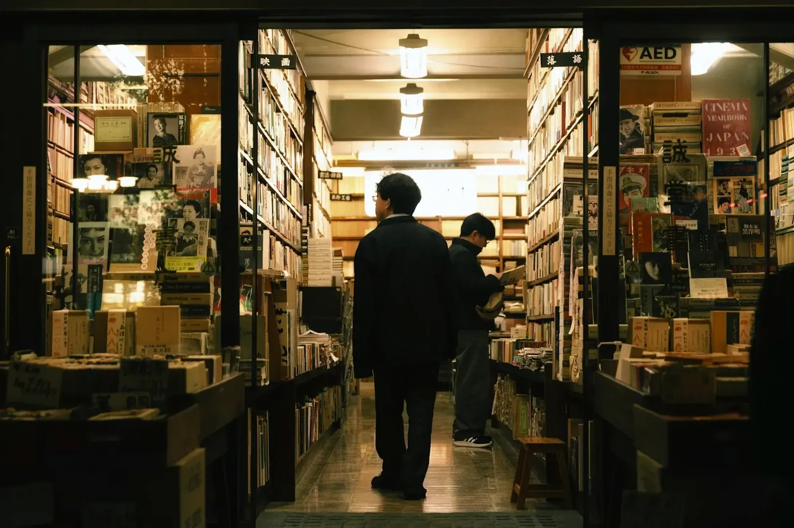 Cozy vintage bookstore with tall shelves filled with books, warm lighting, and customers browsing titles in a narrow aisle.