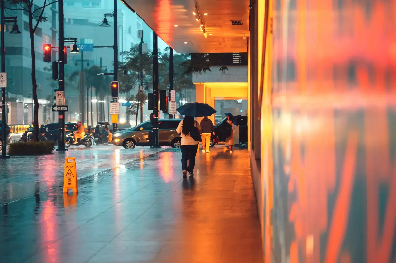 People walking with umbrellas along a wet city street glowing with warm evening lights, portraying how detours and unexpected rain invite a deeper, intimate attention to a place.