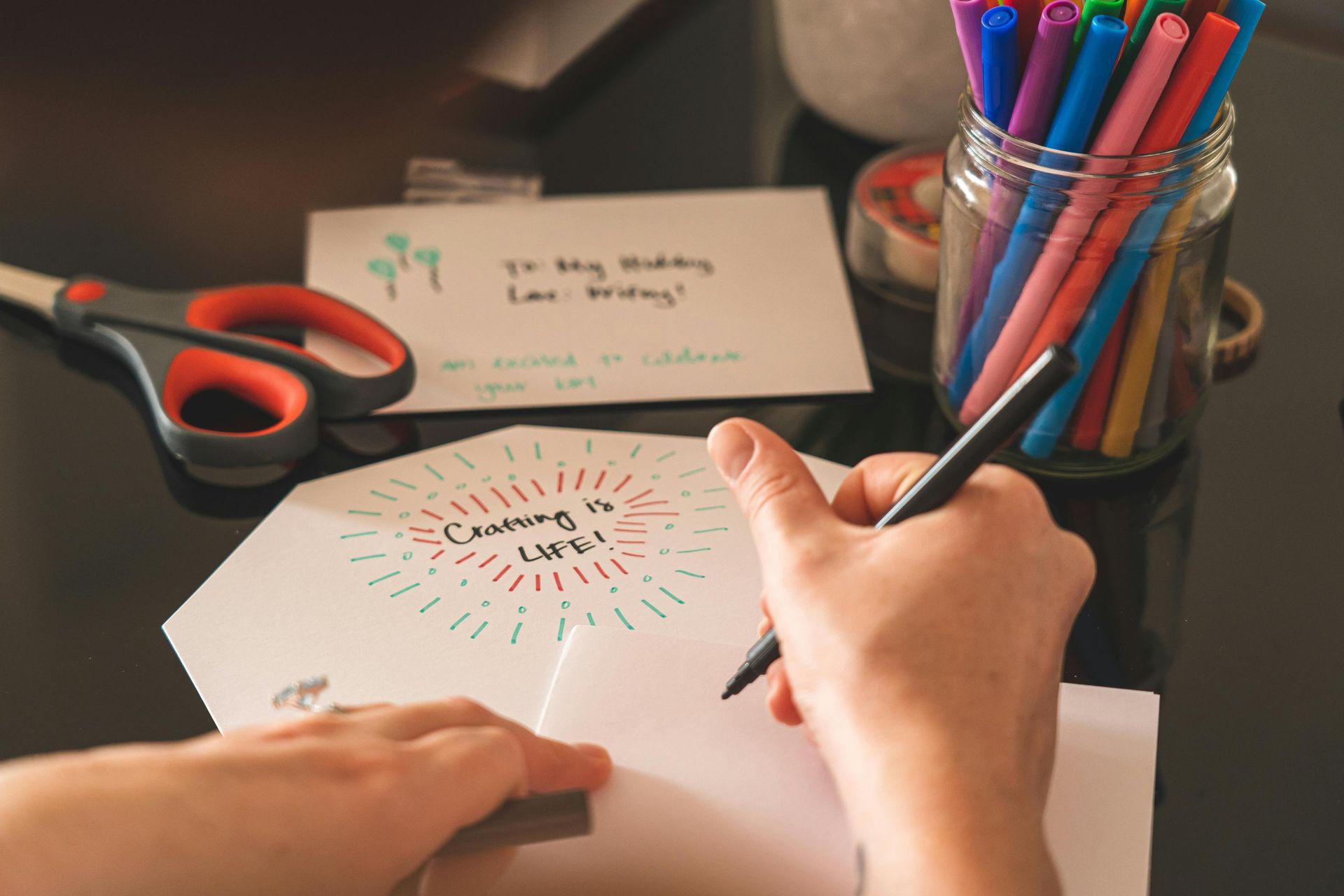 A pair of hands uses a black marker to write on a white card, surrounded by various art supplies including a jar of colorful pens and scissors. A nearby hexagonal note featuring the phrase