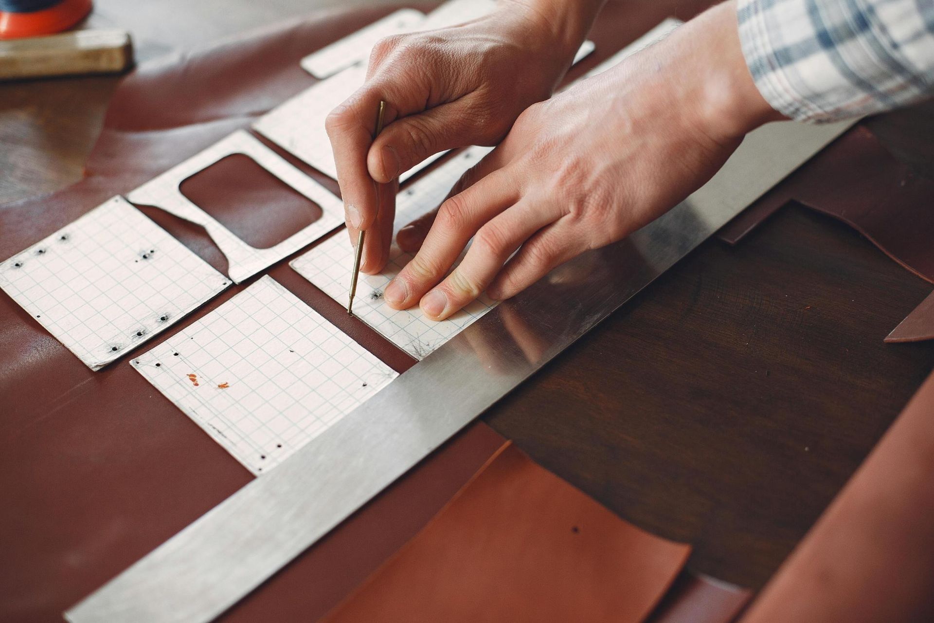 This close-up image shows an artisan's hands carefully working on a sheet of brown leather using a metal stylus and a straight ruler. Several white paper templates featuring grid lines are positioned on the material to guide the precise marking process.