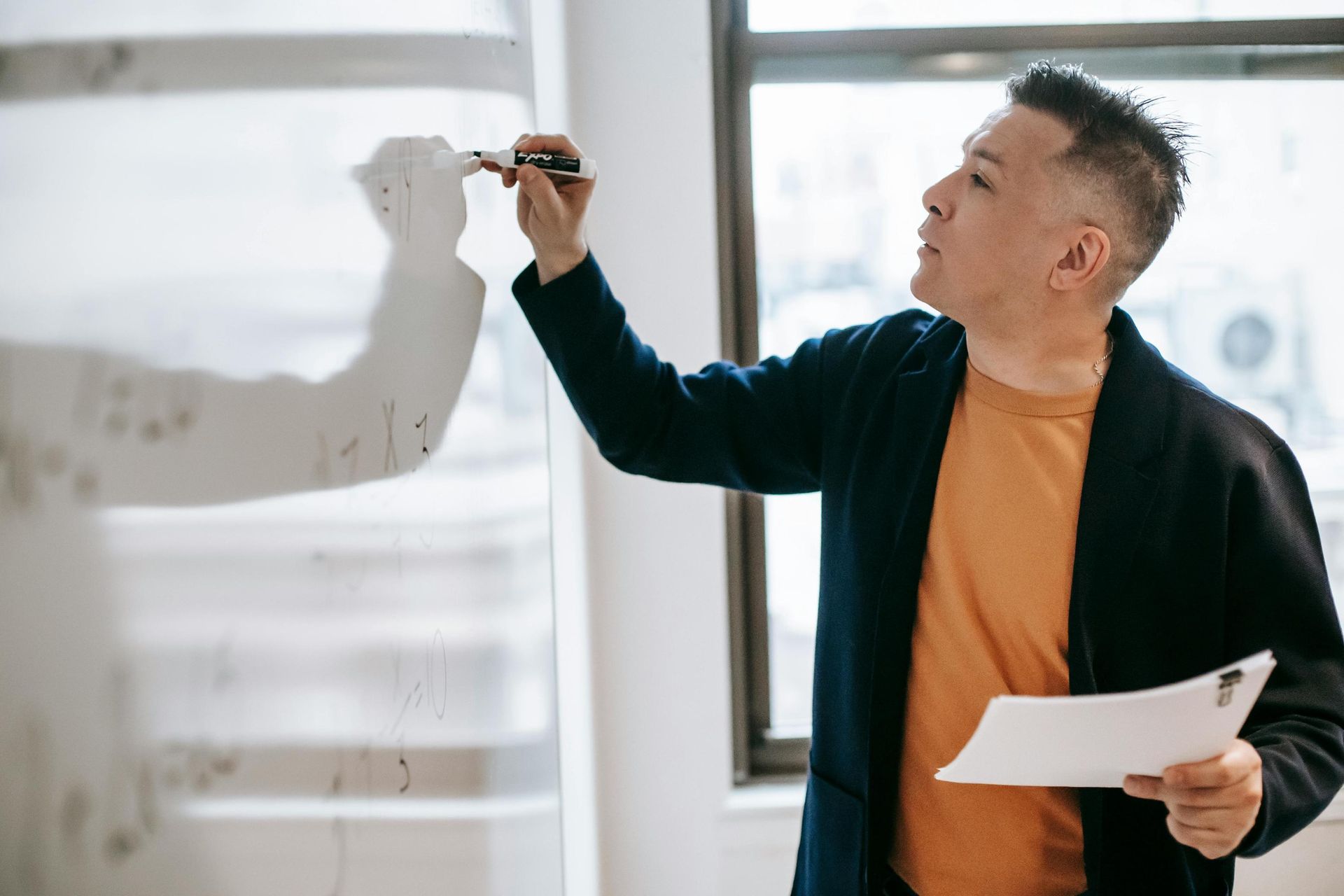 A man in a dark blazer and orange shirt stands before a transparent glass board, focused on writing notes or equations with a marker. He holds a stack of papers in his other hand, appearing deeply engaged in a presentation or a collaborative brainstorming session.