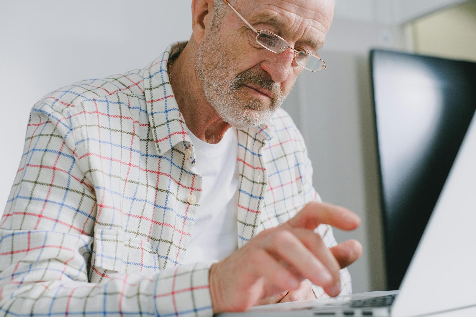 A close-up view captures an elderly, bearded man wearing glasses as he intently focuses on typing on a laptop. He is dressed in a white plaid button-down shirt and appears deep in concentration while working indoors.