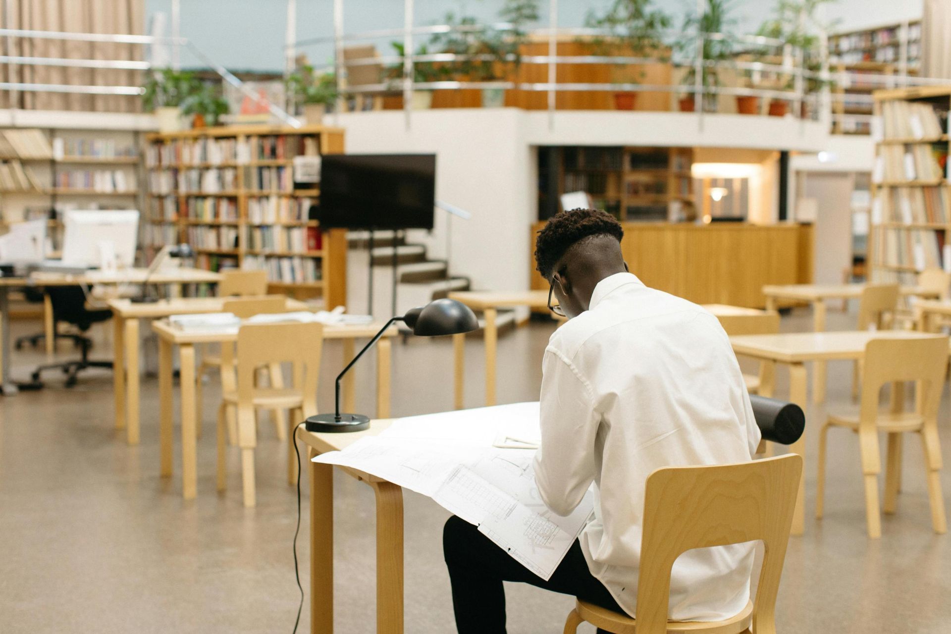 Viewed from behind, a man in a white shirt sits at a wooden desk in a spacious library, intently studying a large document spread out under a desk lamp. The open room is filled with rows of light wooden furniture and bookshelves, featuring a mezzanine level decorated with potted plants.