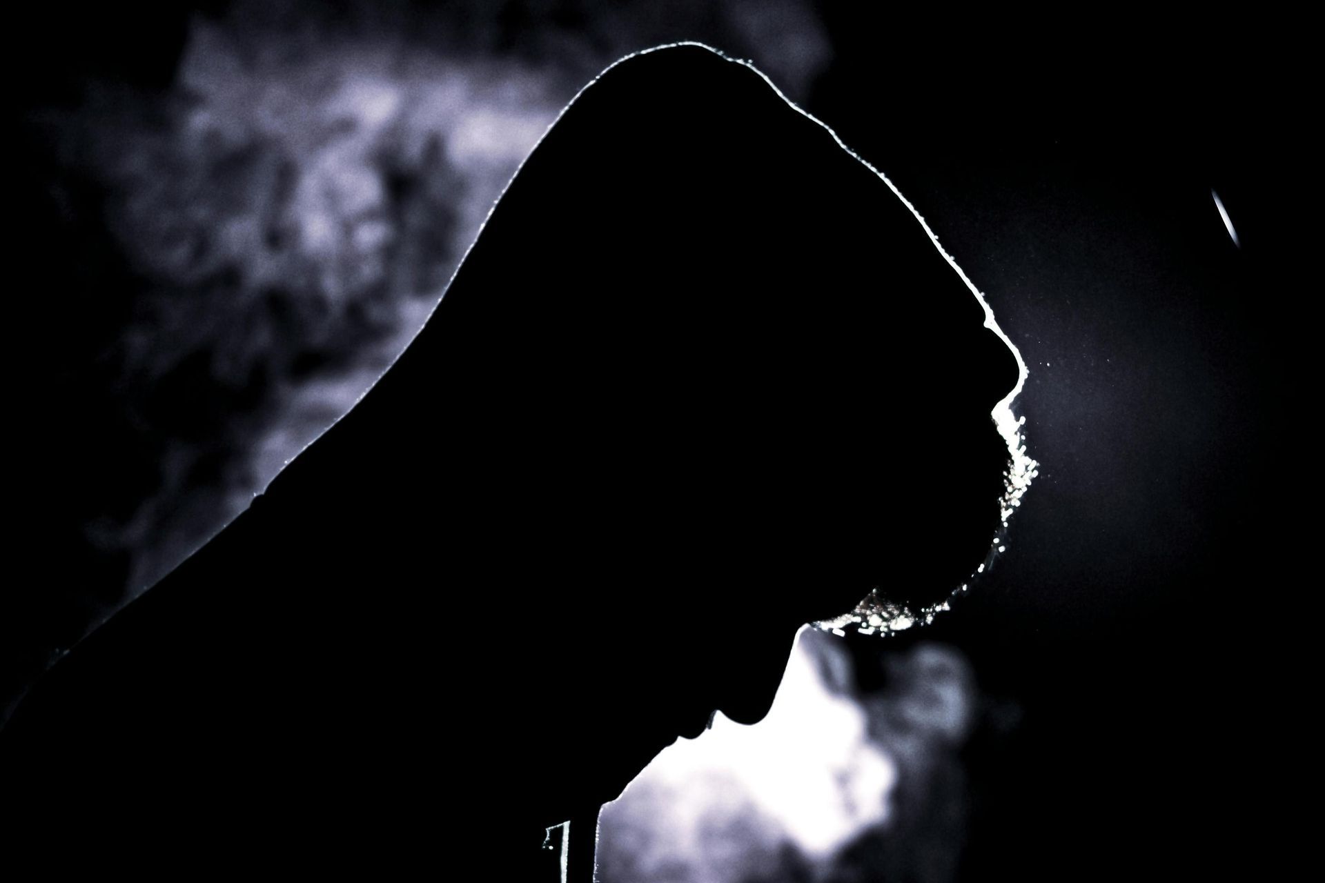 A silhouetted figure wearing a hood is captured in profile against a dark background, with their head tilted downward in a reflective pose. Strong backlighting creates a sharp rim of light along the subject's edge and illuminates a hazy, smoke-like texture swirling behind them.