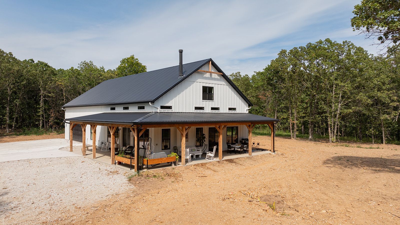 A white house with a black roof and a porch in a field surrounded by trees.