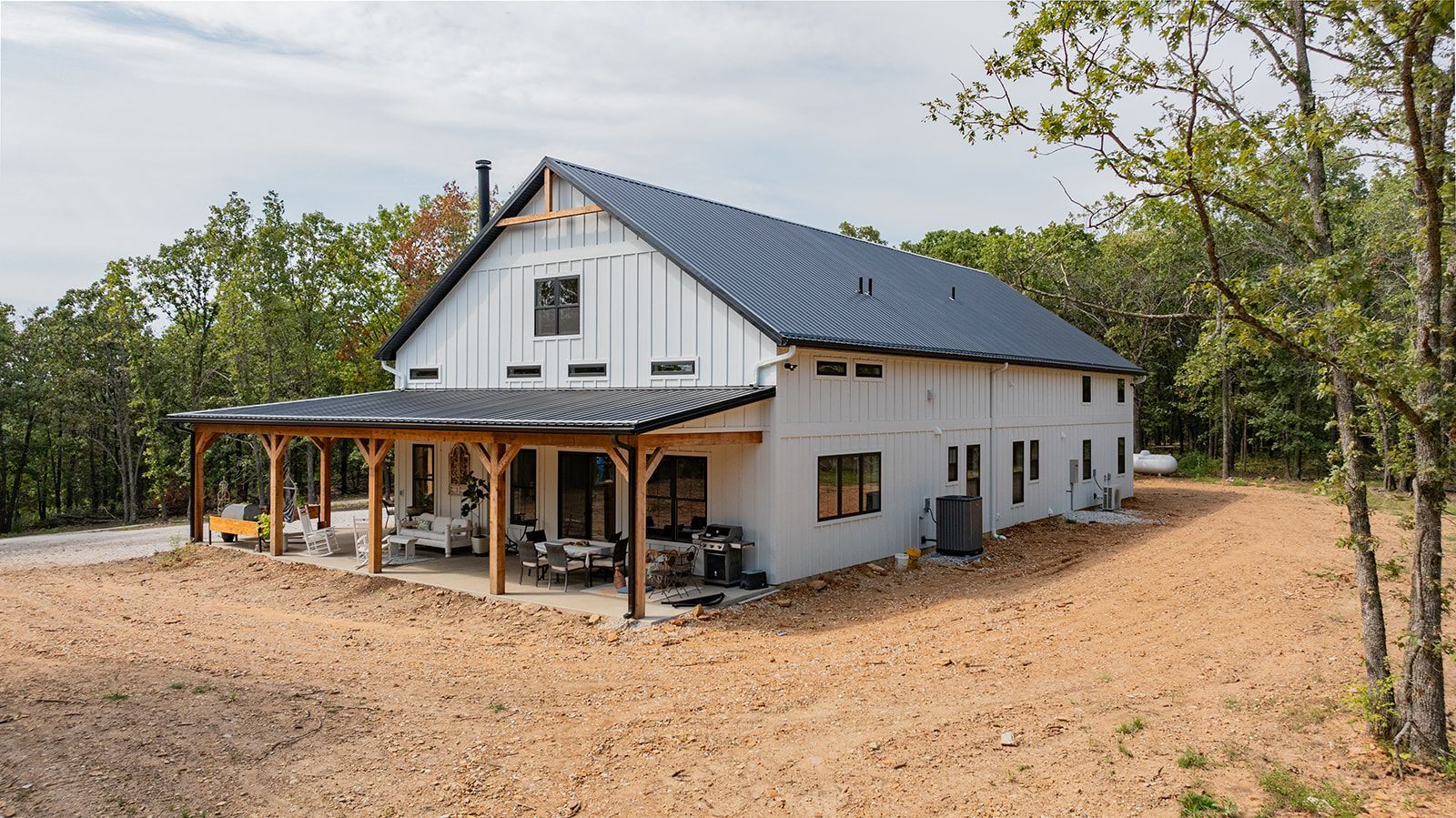 A large white barn with a porch in the middle of a field surrounded by trees.