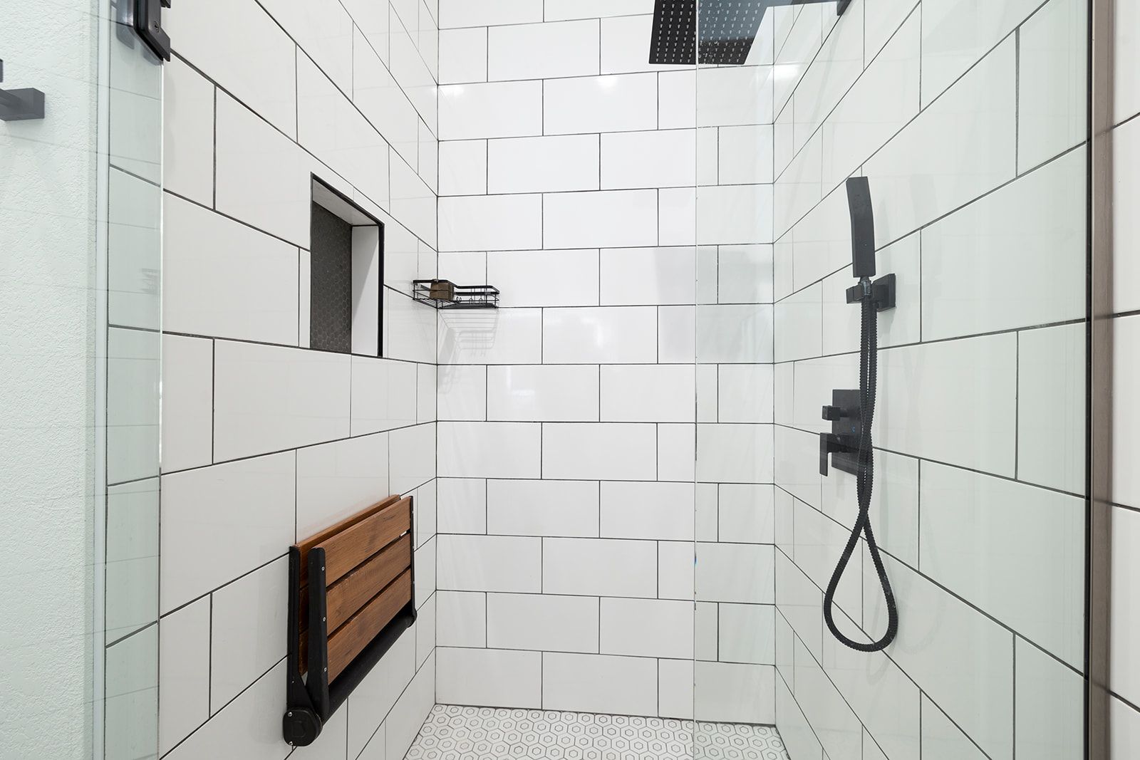 A bathroom with white tiles and a walk in shower with a wooden seat.