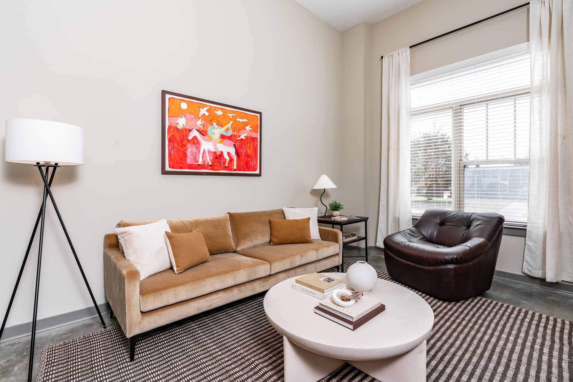Living room with tan sofa, brown armchair, a white coffee table with books and a lamp, and art with a white horse.