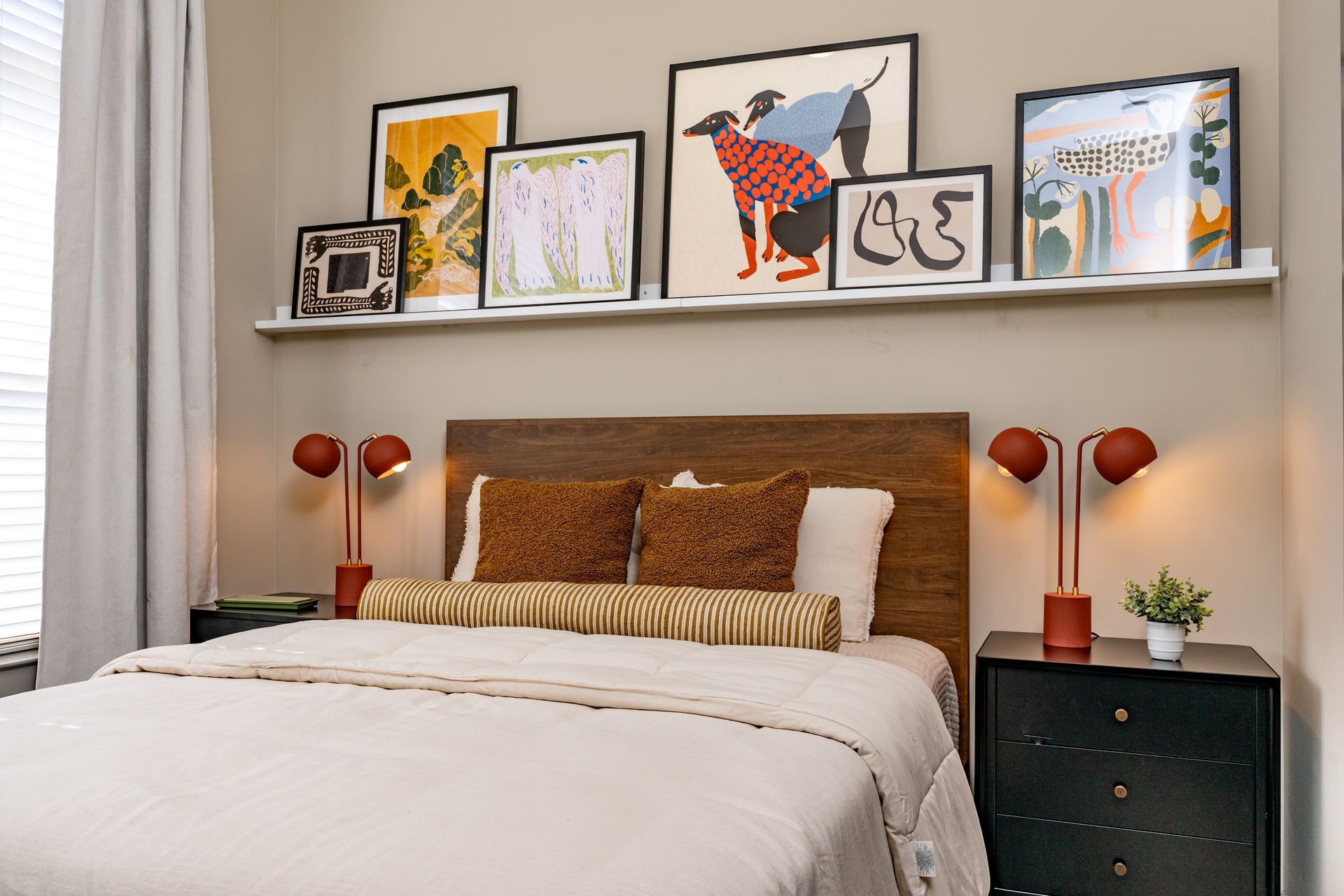 Bedroom with a bed, bedside tables, and artwork on a shelf above. The bed has brown pillows, a white comforter, and wooden headboard.
