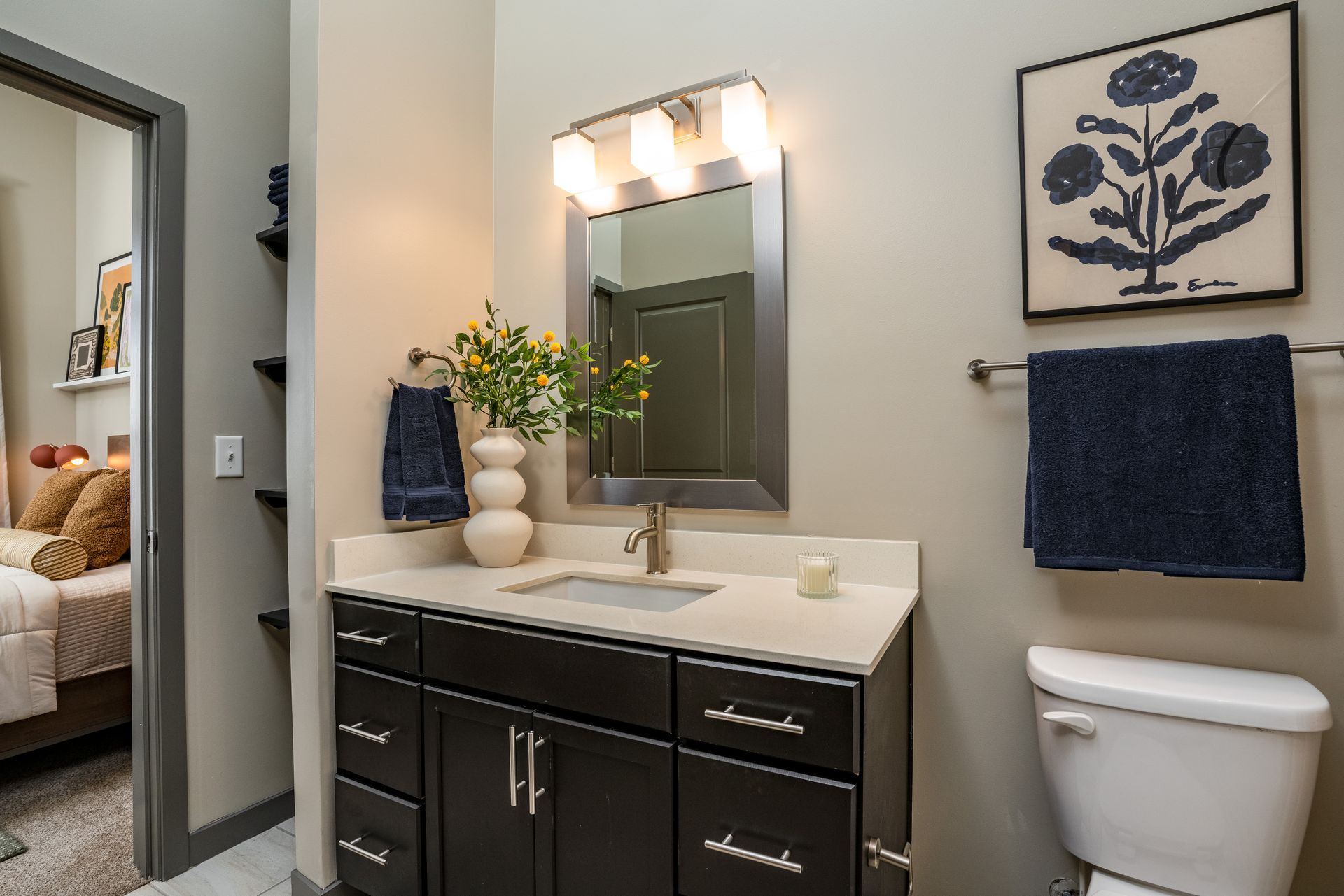 Bathroom with dark wood vanity, white countertop, silver mirror, and floral artwork; a blue towel hangs nearby.