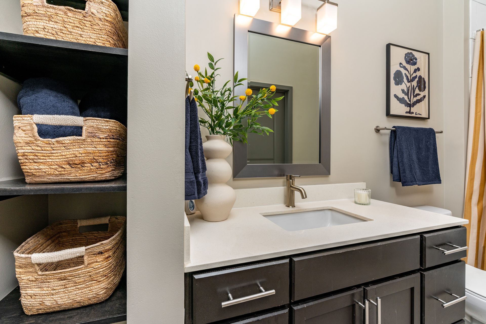 Bathroom with dark cabinets, white countertop, and wicker baskets on black shelves. A blue towel hangs on the wall next to a framed flower print.