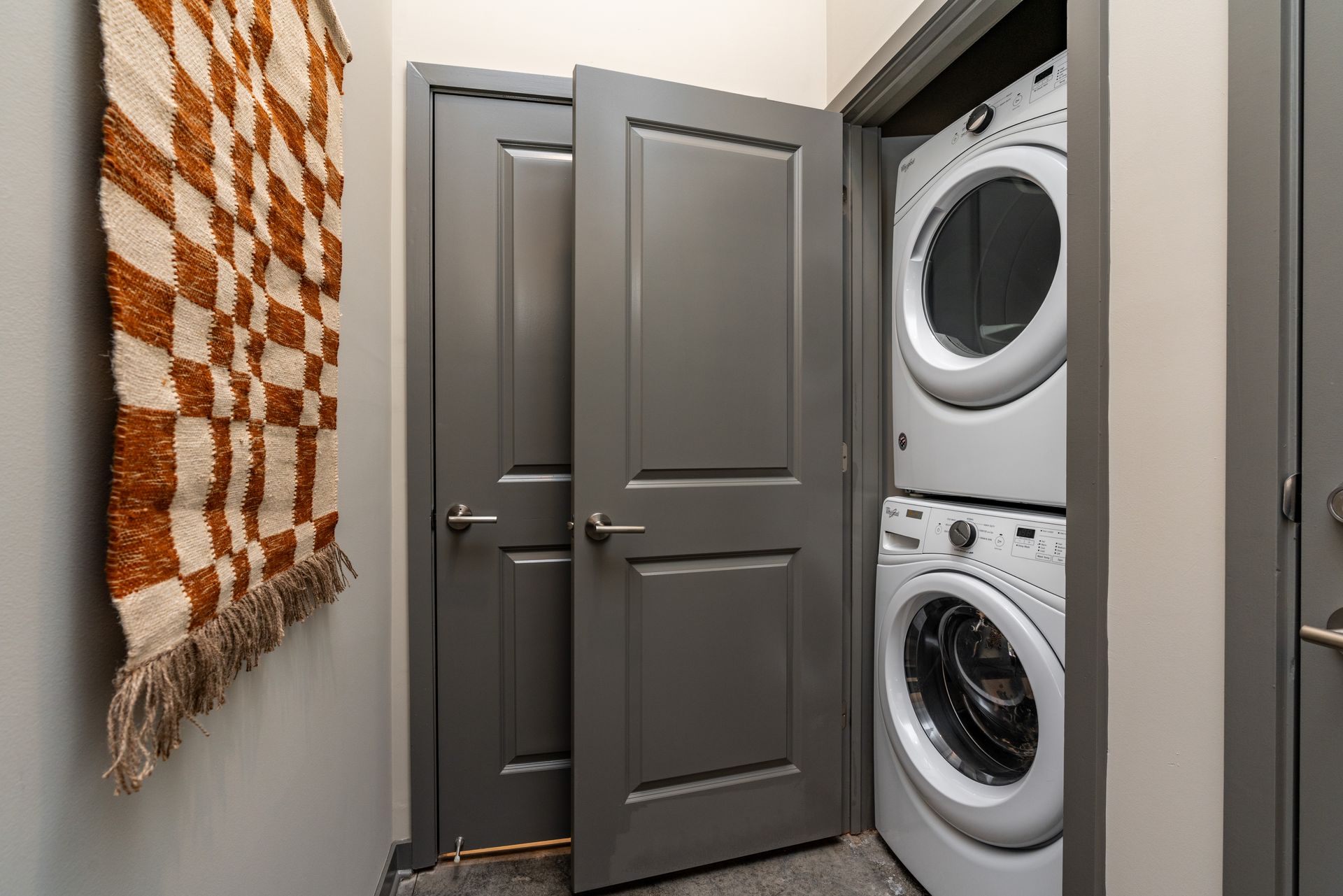 Laundry room with stacked washer and dryer next to a closed gray door. A woven wall hanging is on the left.
