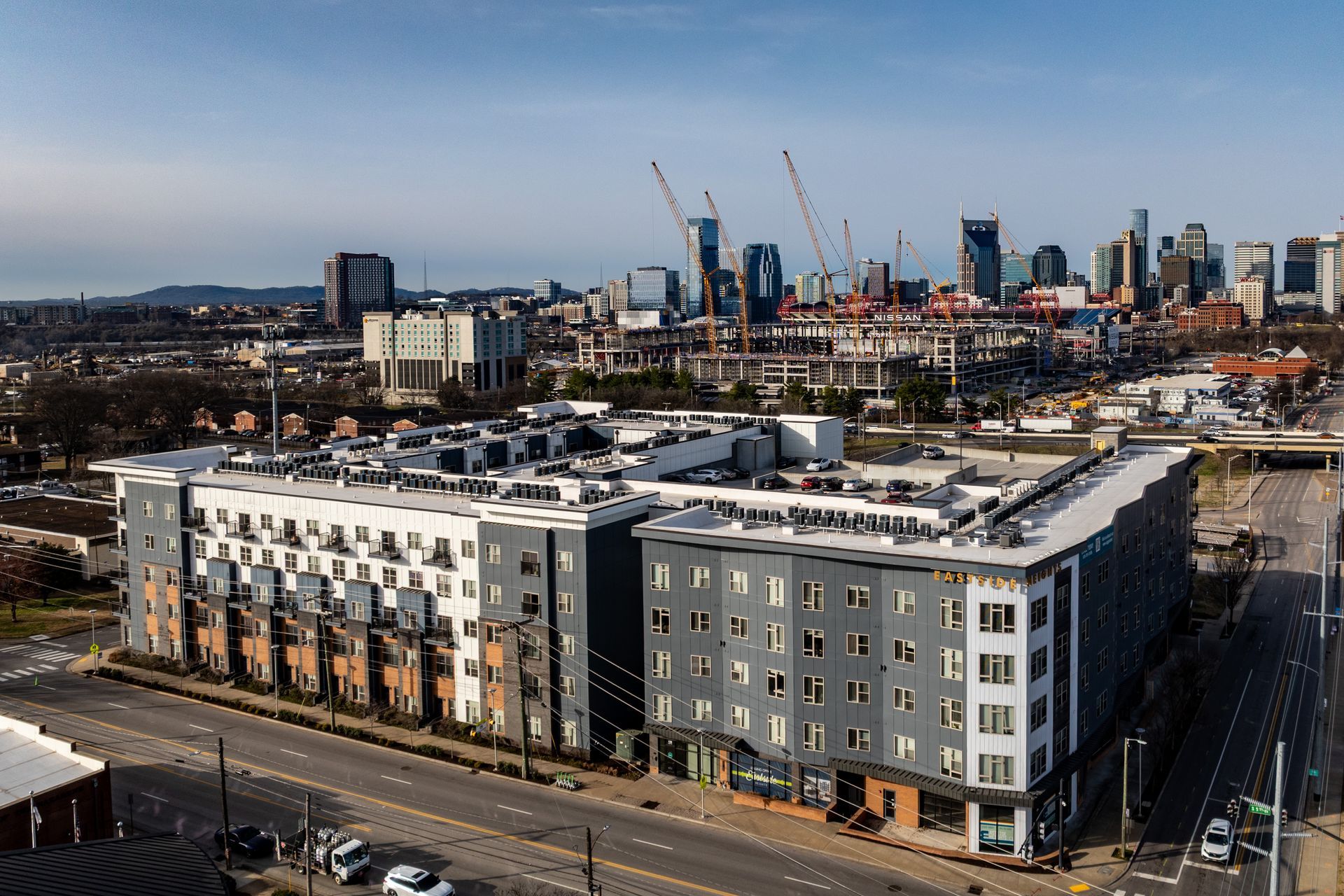 Aerial view of modern apartment buildings in front of the Nashville skyline with construction cranes. Gray and white buildings line a city street.