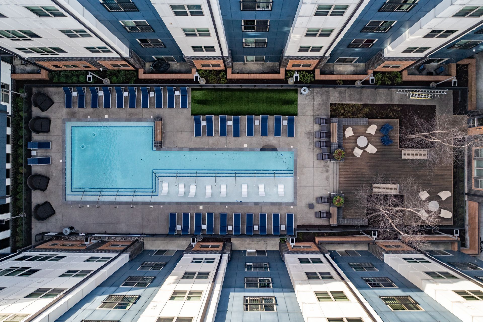Aerial view of an apartment complex courtyard with a pool, lounge chairs, and a wooden patio with seating. Blue and white buildings surround the area.