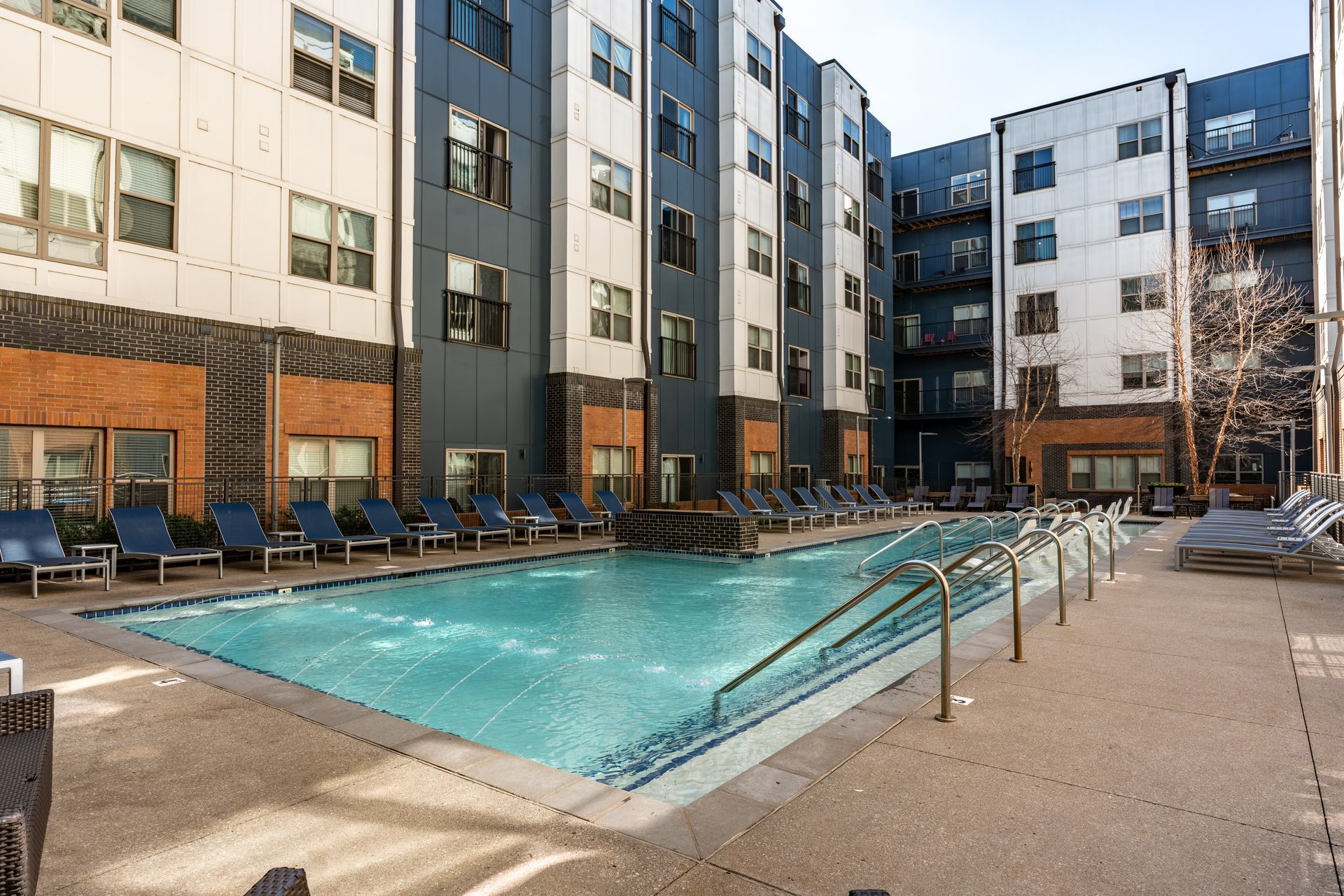 A rectangular pool surrounded by lounge chairs and a multi-story apartment building with blue and brick walls.