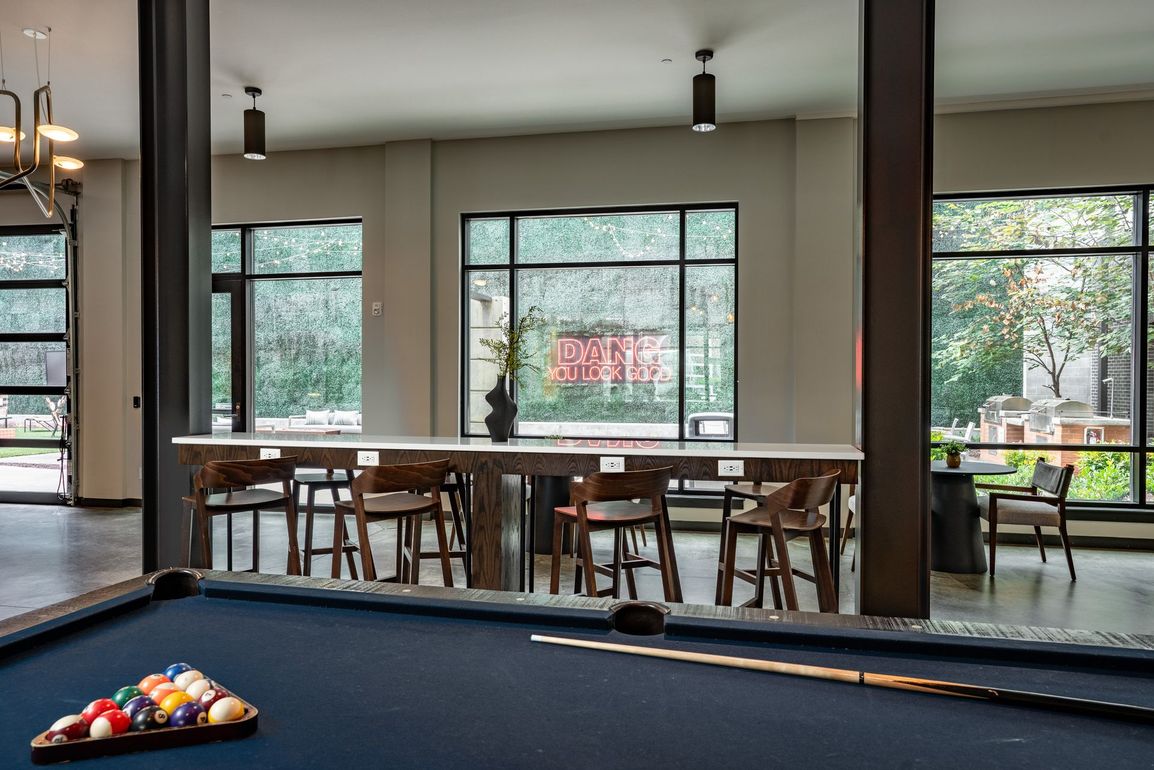 A pool table in a modern lounge with a long table, chairs, and large windows overlooking greenery.