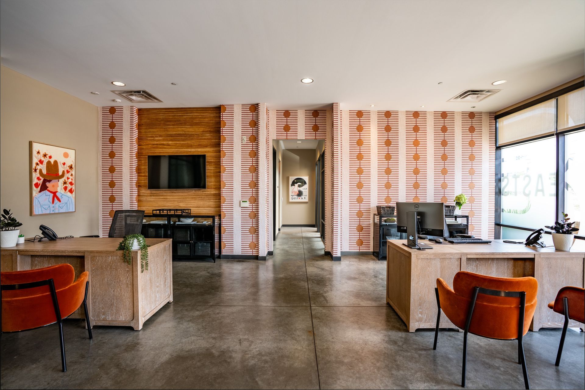 Modern office interior with desks, orange chairs, and patterned wallpaper. The space features a TV and artwork.