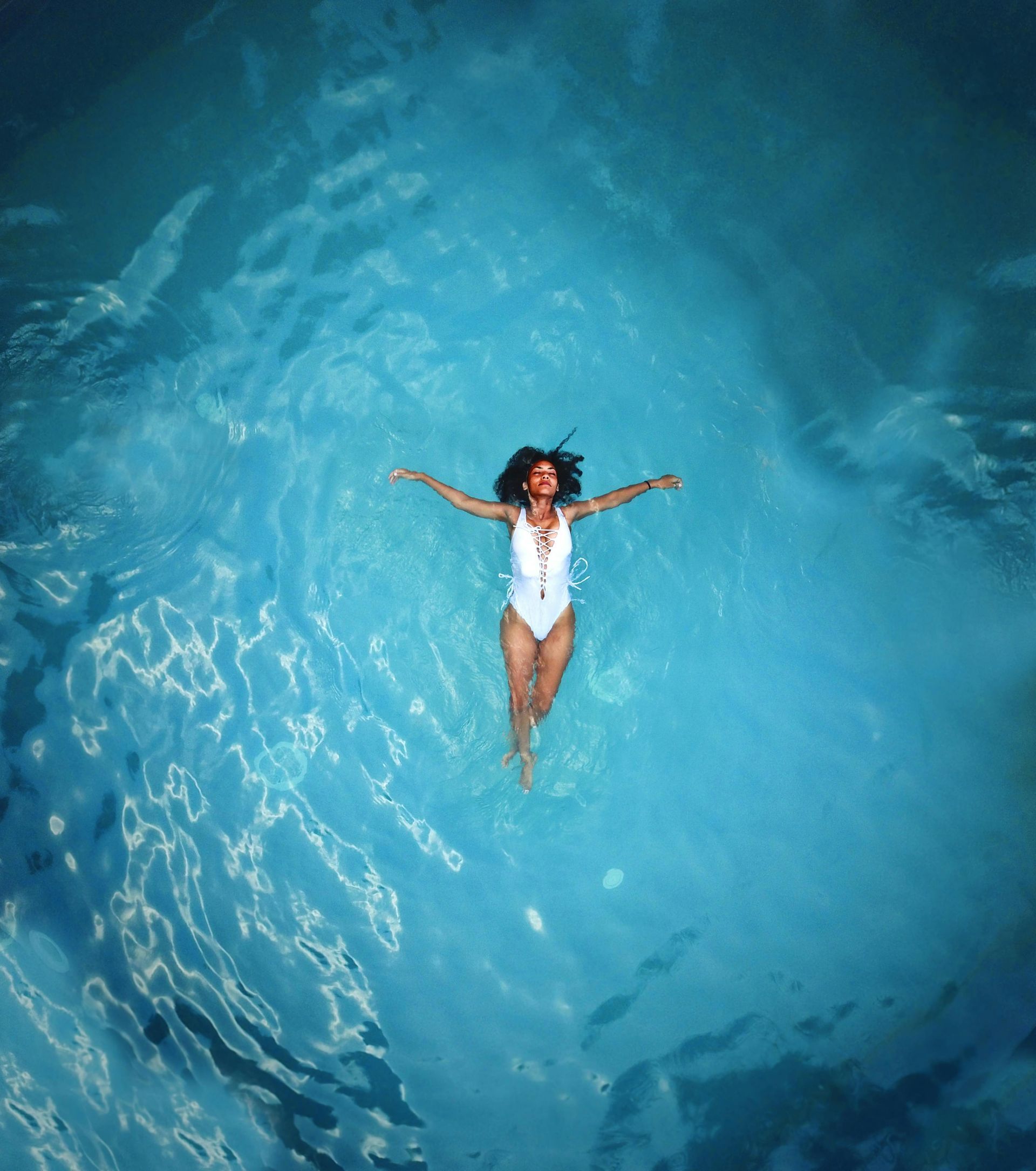 Woman in white swimsuit floating in a bright blue pool, arms outstretched, viewed from above.