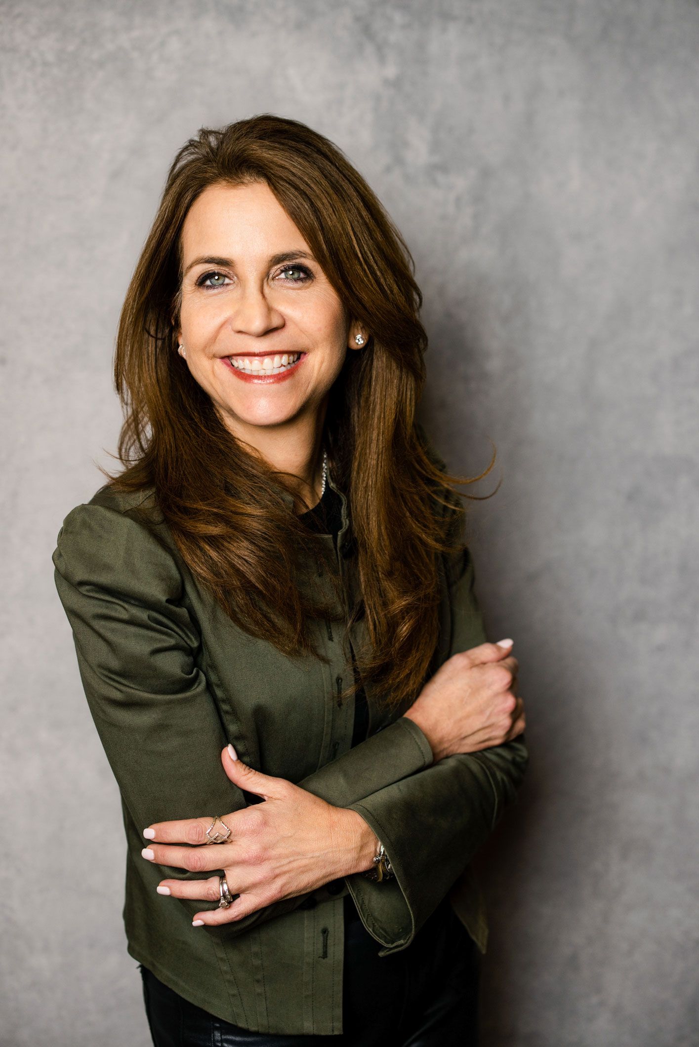 A smiling woman with long brown hair, wearing a dark olive jacket, standing against a grey textured background. A smiling woman with long brown hair, wearing a dark olive jacket, standing against a grey textured background.