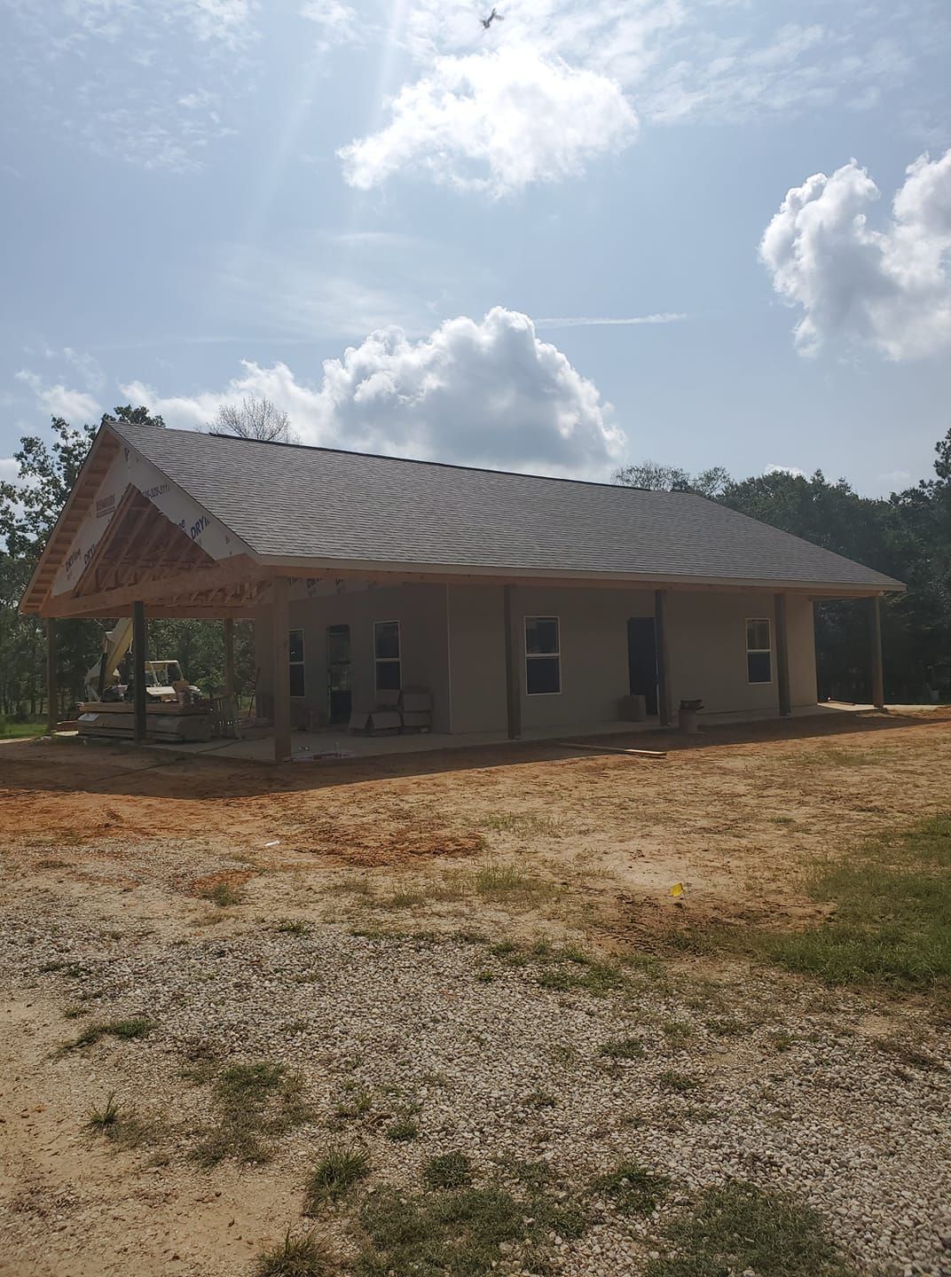 Single-story house with a sloped roof under a bright cloudy sky, surrounded by a gravel yard.
