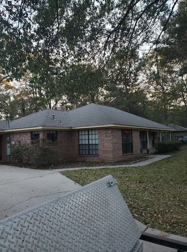 Single-story brick house with a gray roof and driveway, seen through trees at dusk.