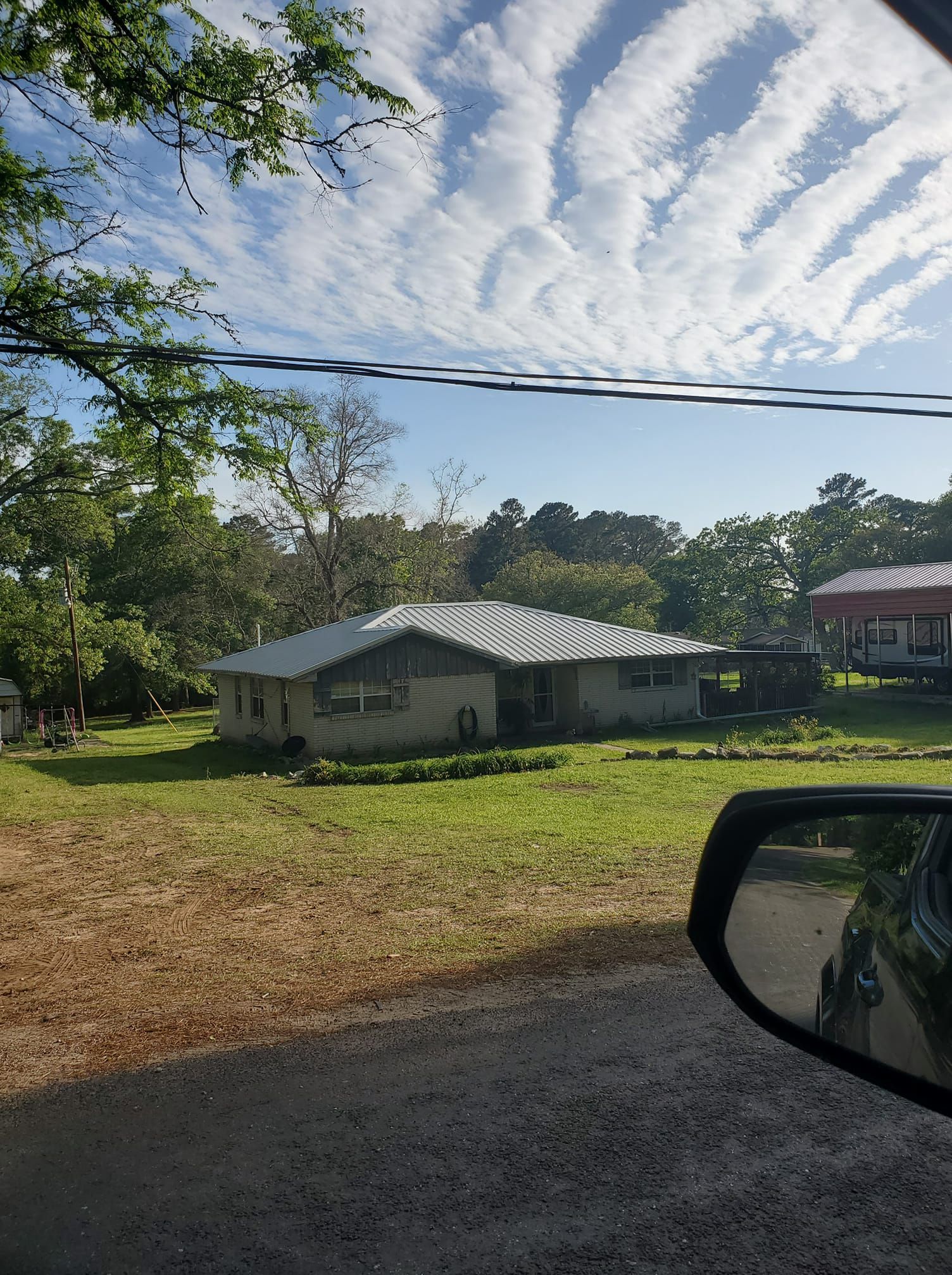 Rural roadside view with a small house, green yard, power lines, and a cloudy blue sky.