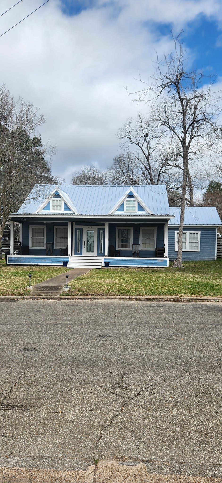 Blue house with white trim and front porch on a gravel driveway under a cloudy sky