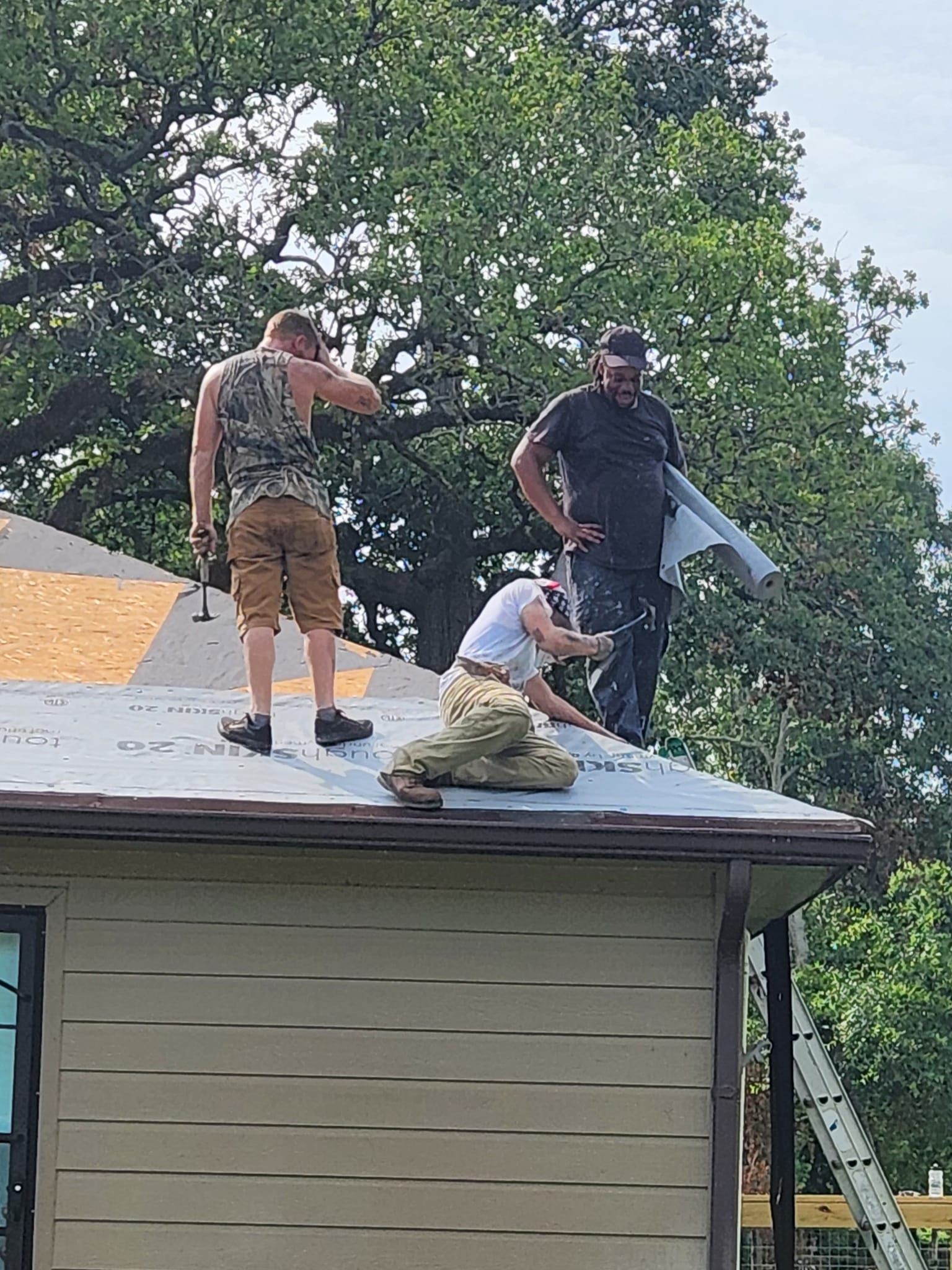 Three people working on a roof, handling shingles and tools near a chimney.