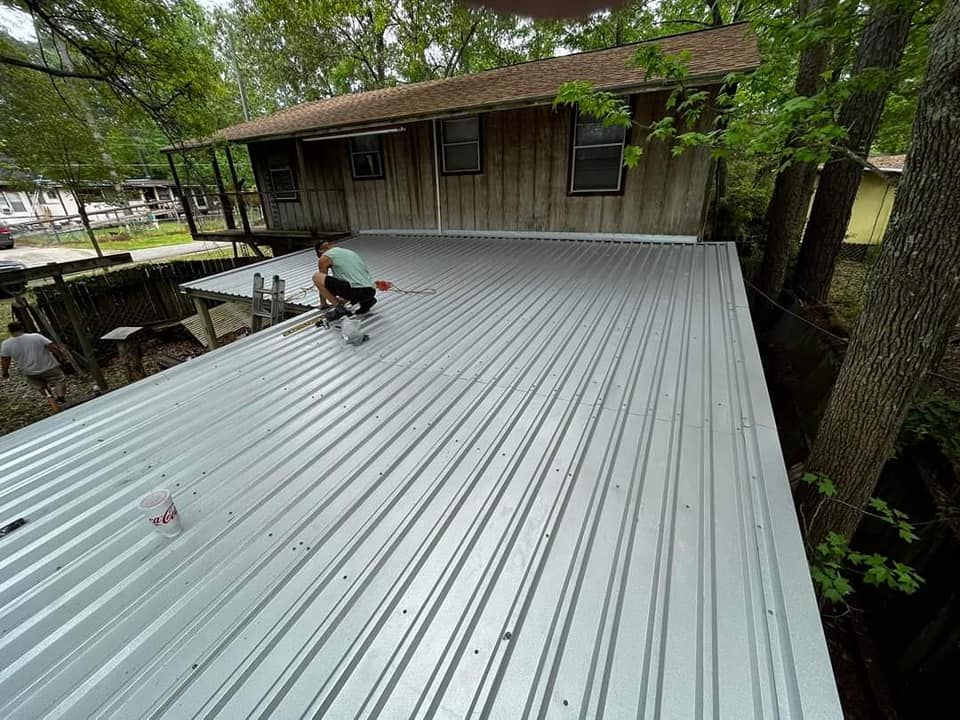 Worker painting a gray corrugated metal roof beside a wooden house surrounded by trees