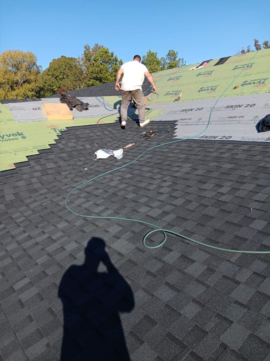 Worker installing roofing shingles on a sloped roof with tools and a green hose.