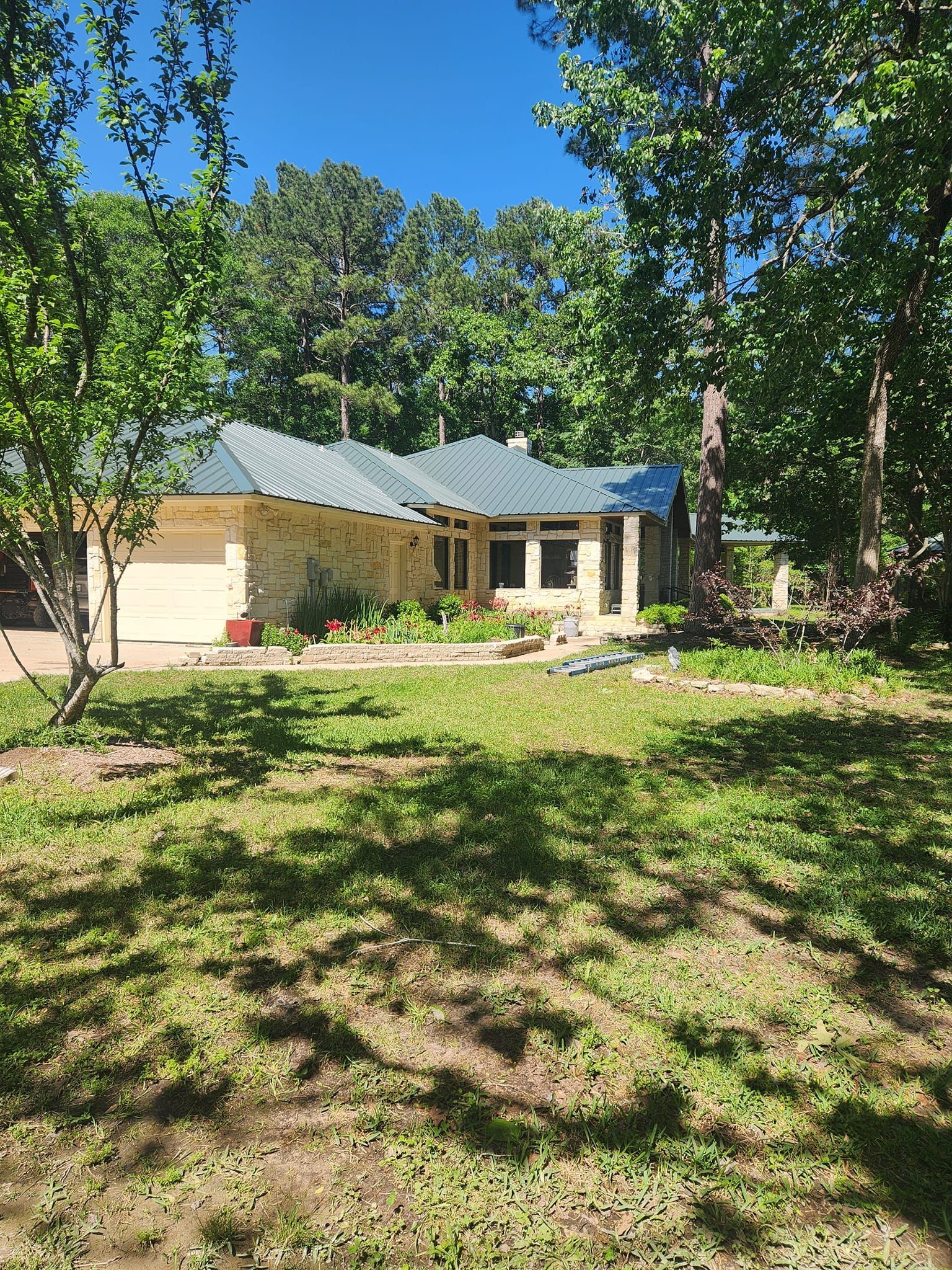 Single-story house with tan siding and gray roof, surrounded by trees and a sunny front yard.