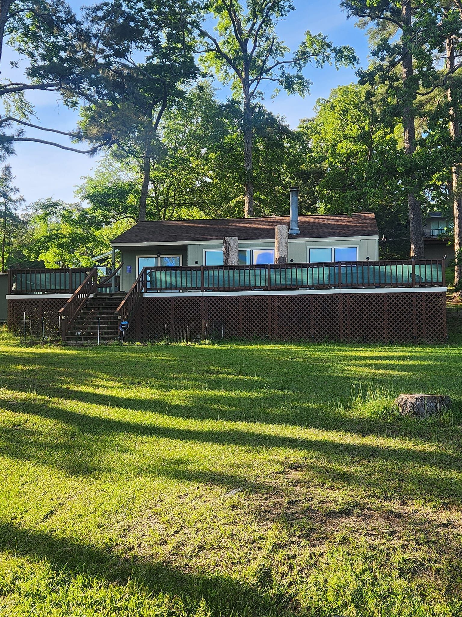 Wooden house with a deck and patio chairs behind a grassy yard and tall trees