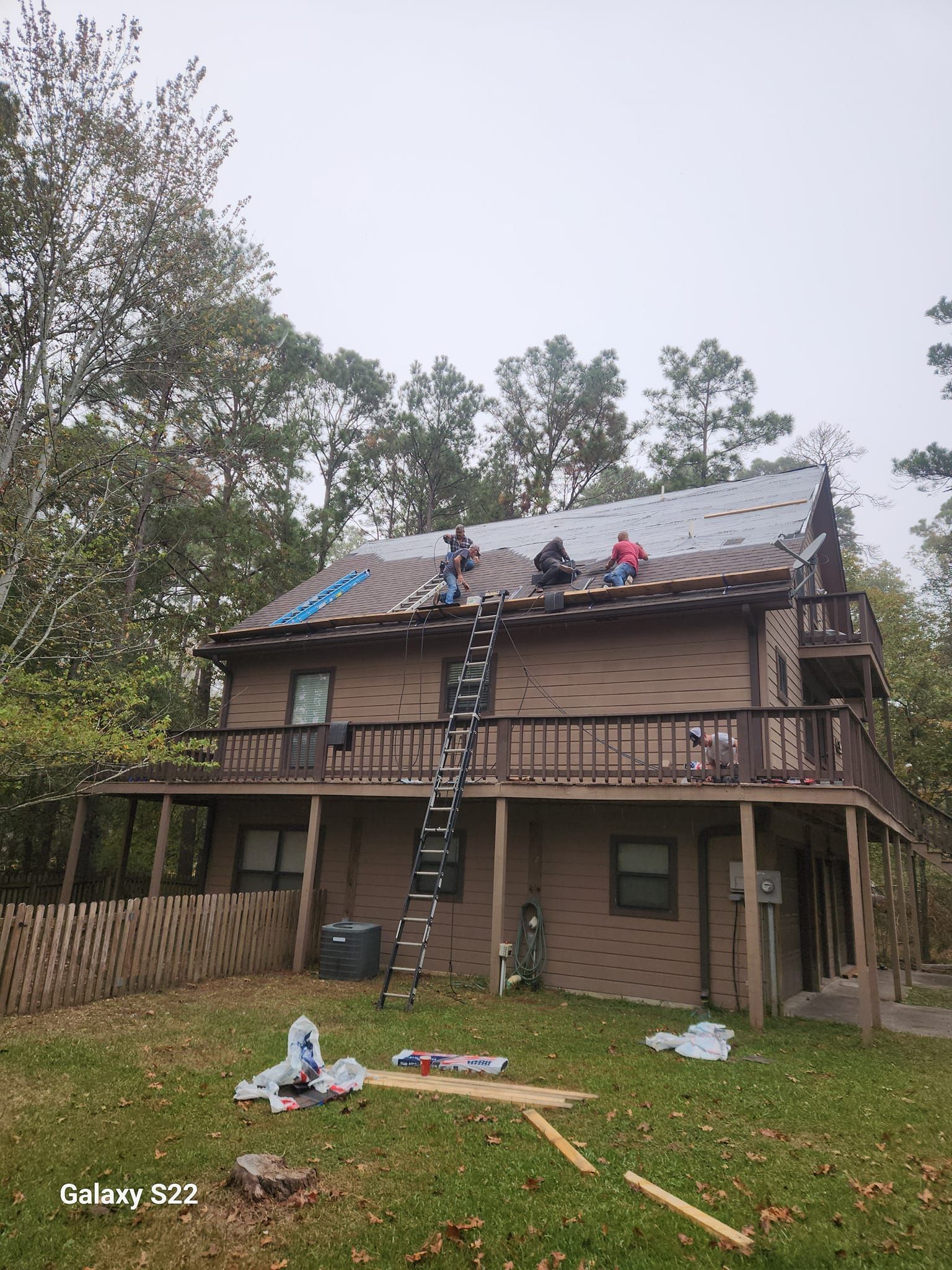 Two-story house with workers repairing the roof, ladder on the front, and tools scattered in the yard.