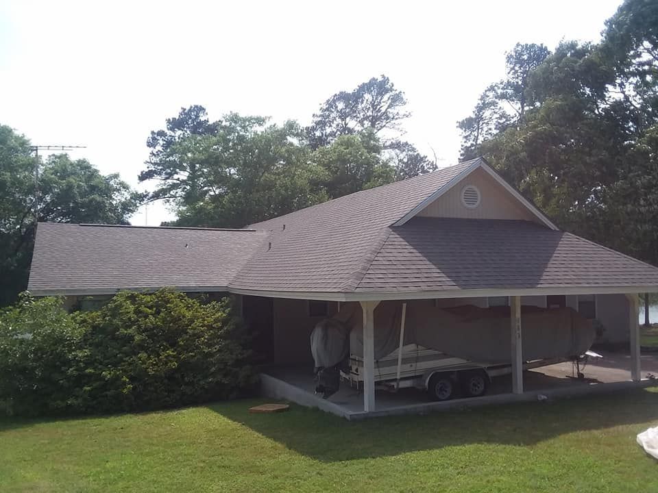 Carport with a brown shingled roof, white posts, and a car parked underneath, surrounded by trees.