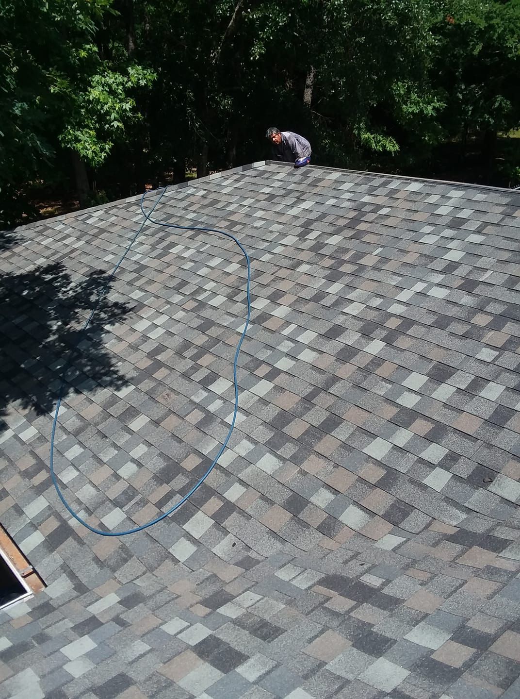 Person working on a shingled roof with a blue hose, surrounded by trees.