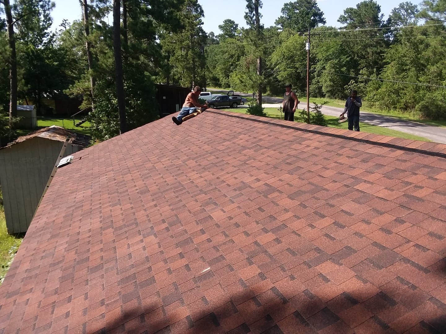 Workers on a shingled roof near a wooded roadside, one using a tool and others standing nearby.