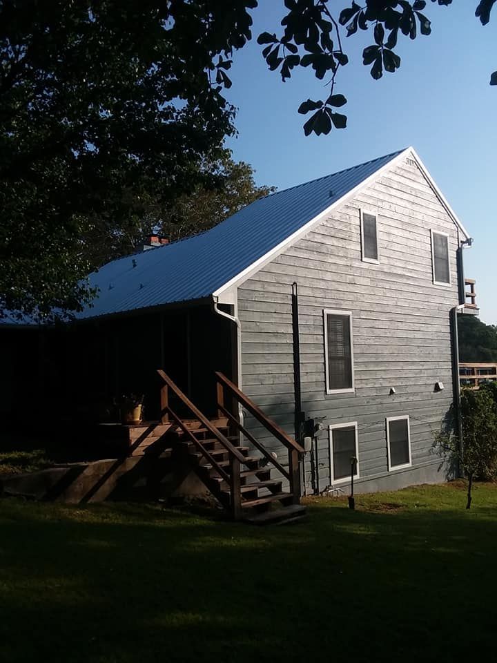 Gray house with a blue metal roof, front porch, and wooden stairs, surrounded by trees and lawn.