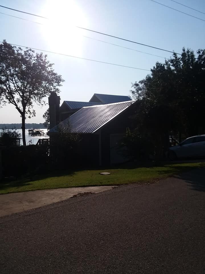 Sunlit house with metal roof by the water, framed by trees and a road in the foreground