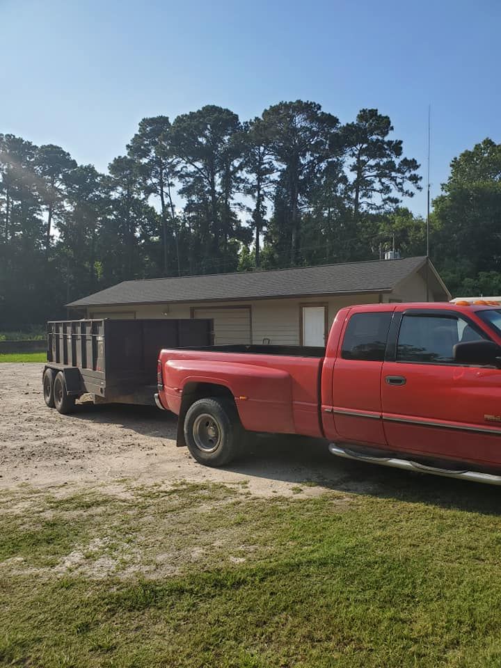Red pickup truck pulling a trailer on a dirt lot beside a small building and trees.